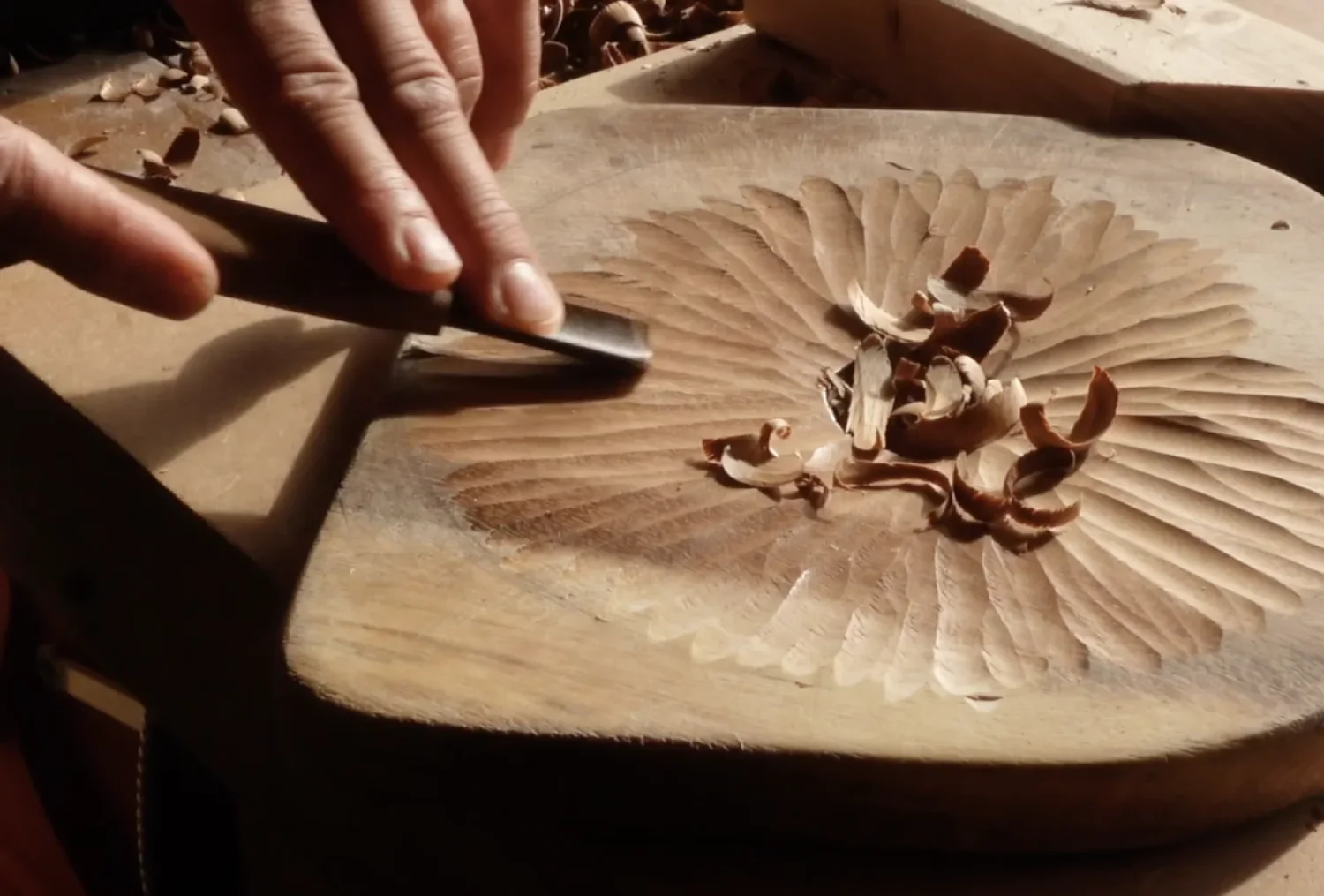 A person is carving a design into a wooden surface with a chisel, surrounded by wood shavings.