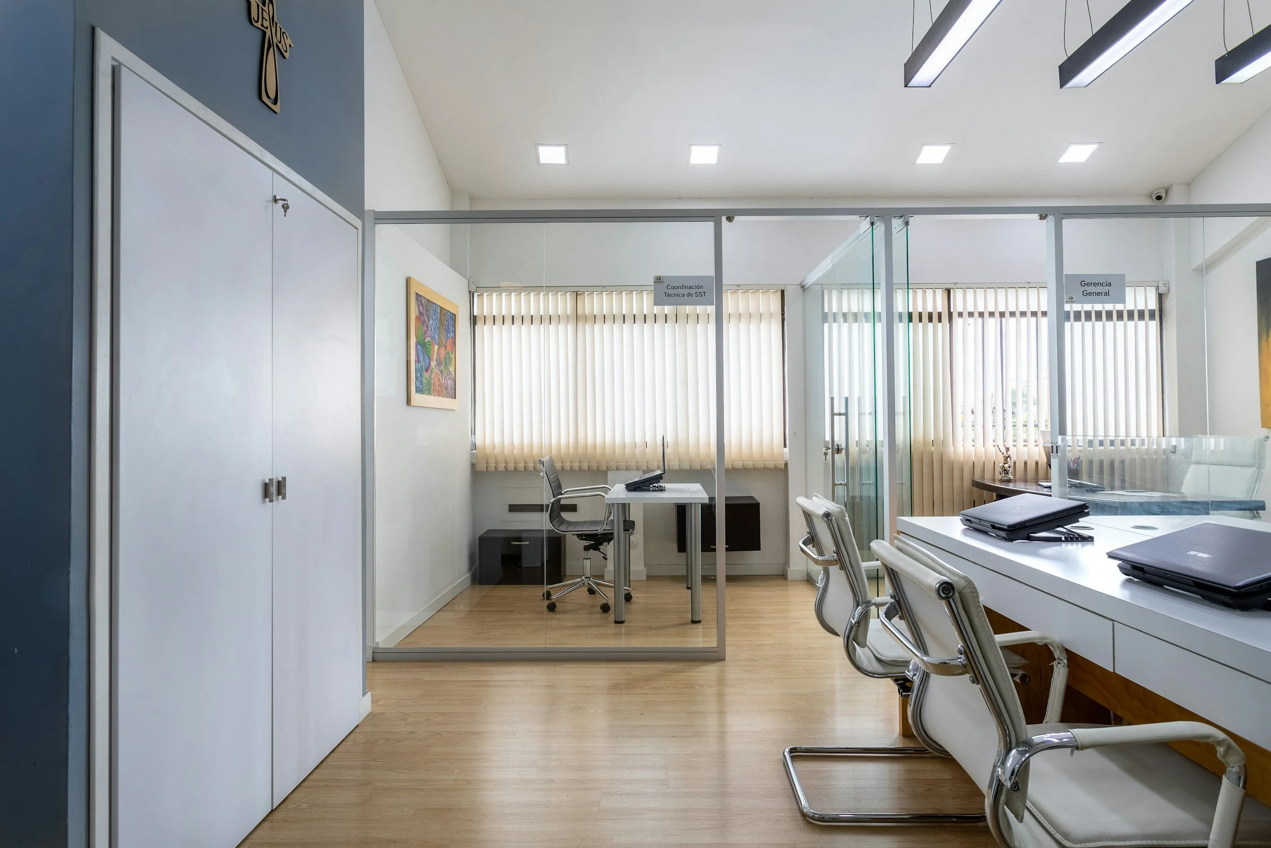 An office space with white walls and wooden flooring, divided by glass partitions. There are white desks and office chairs, with closed laptops on the desks. A colorful painting hangs on the wall, and there are window blinds allowing natural light. Signs indicate office areas, one labeled 'Coordinación Técnica SST' and another 'Gerencia General.'