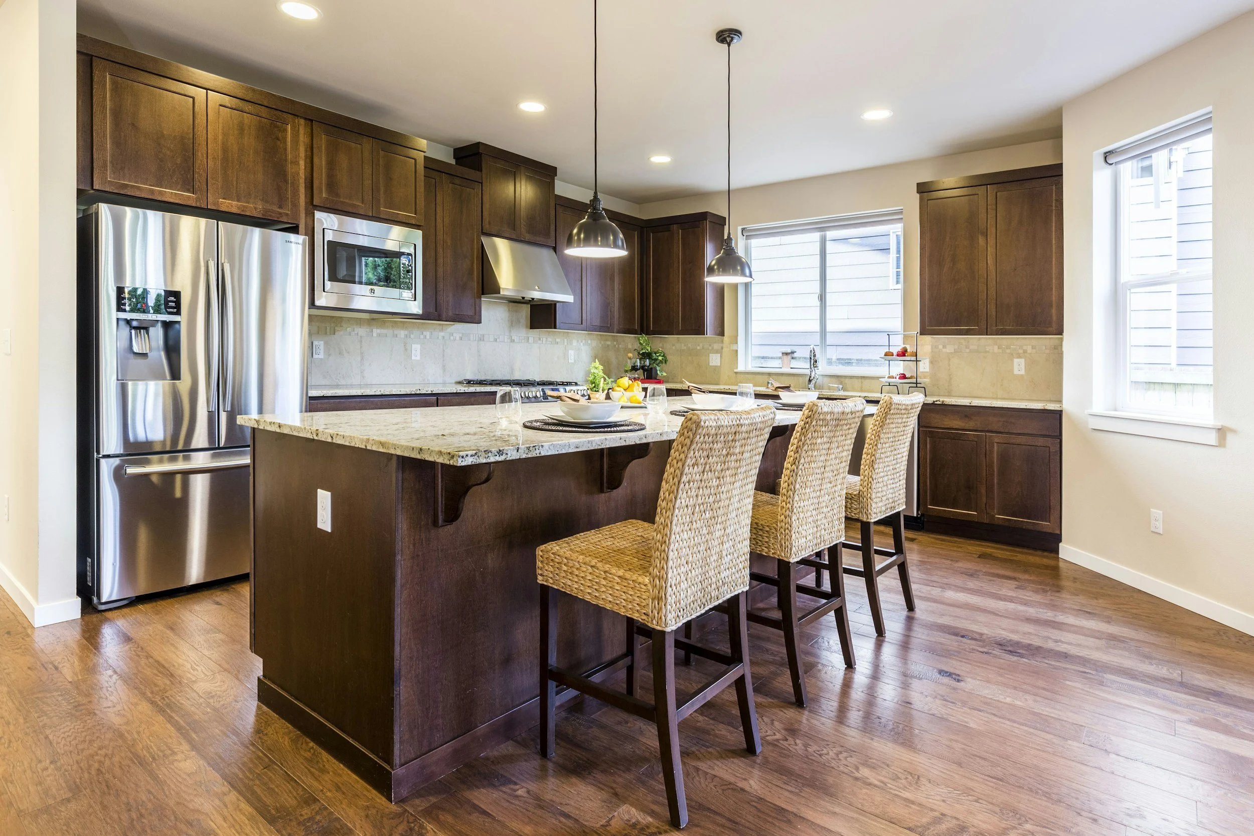 Kitchen with dark wood cabinets, stainless steel appliances including a refrigerator and microwave, granite island with four woven rope bar stools, beige tile backsplash, and two hanging pendant lights. Windows with white blinds provide natural light.