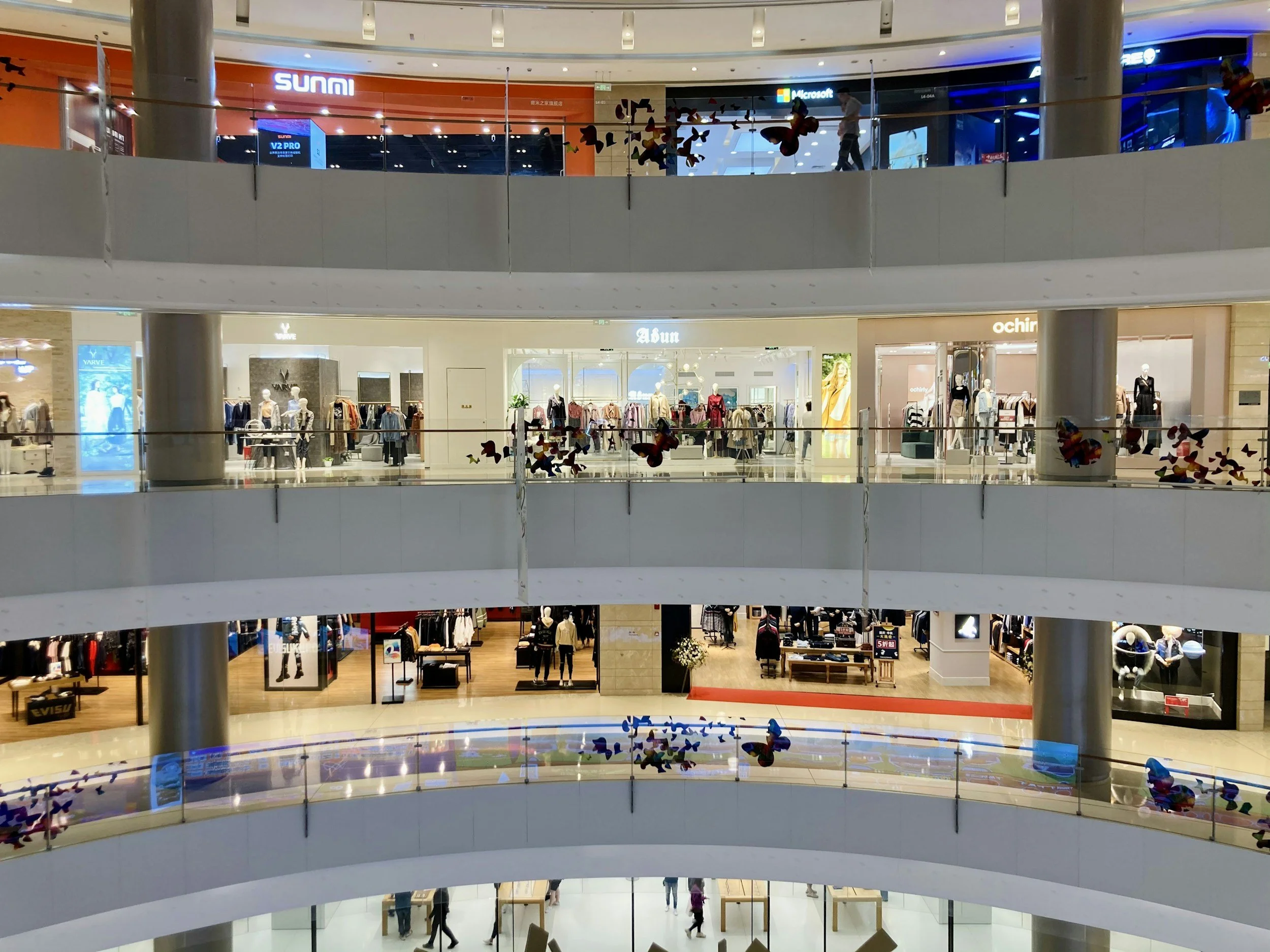 Interior of a multi-level shopping mall with various clothing stores, mannequins dressed in outfits, and shoppers walking.