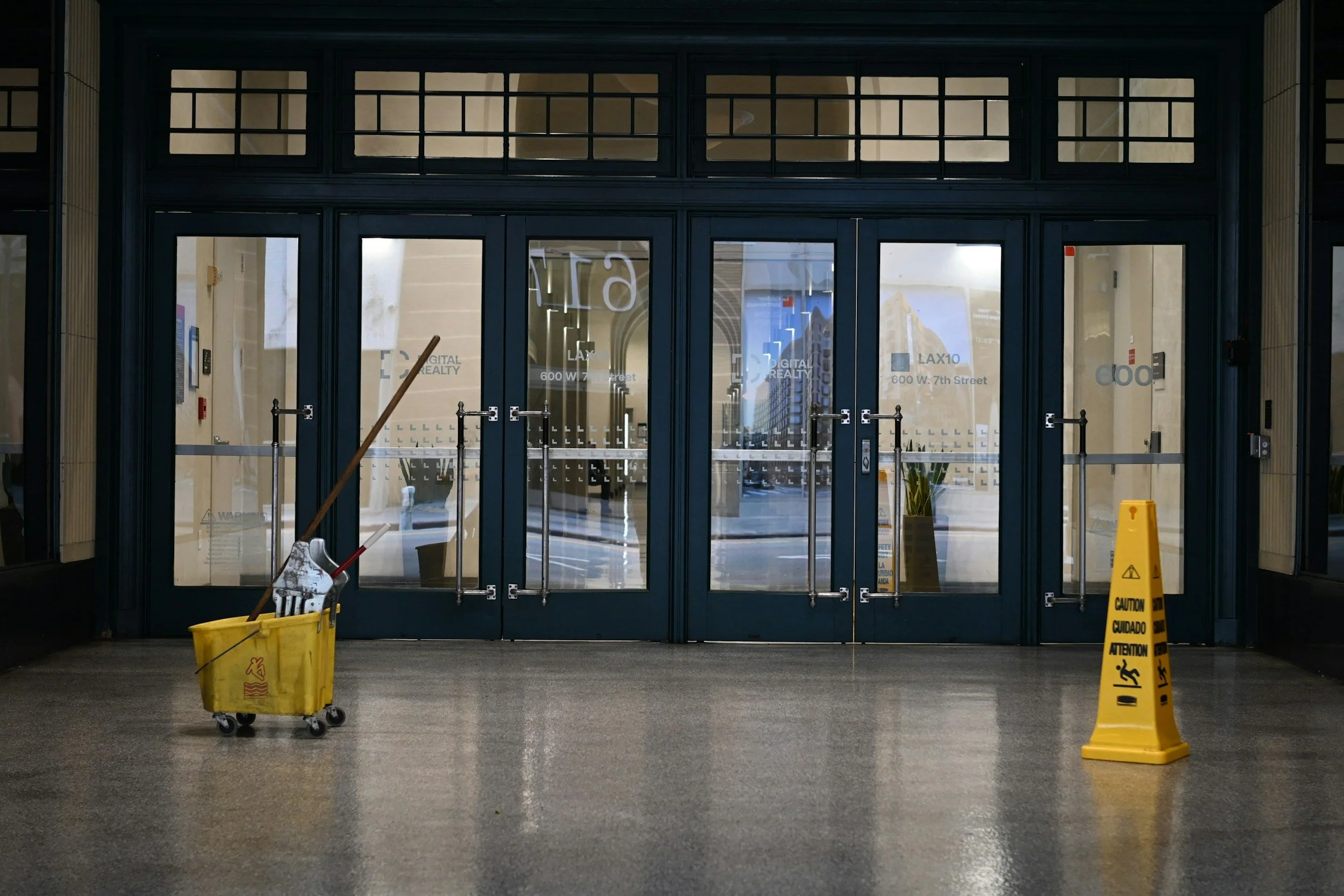 Front entrance of a building with glass doors, cleaning cart, yellow caution signs, and floor cleaning equipment.
