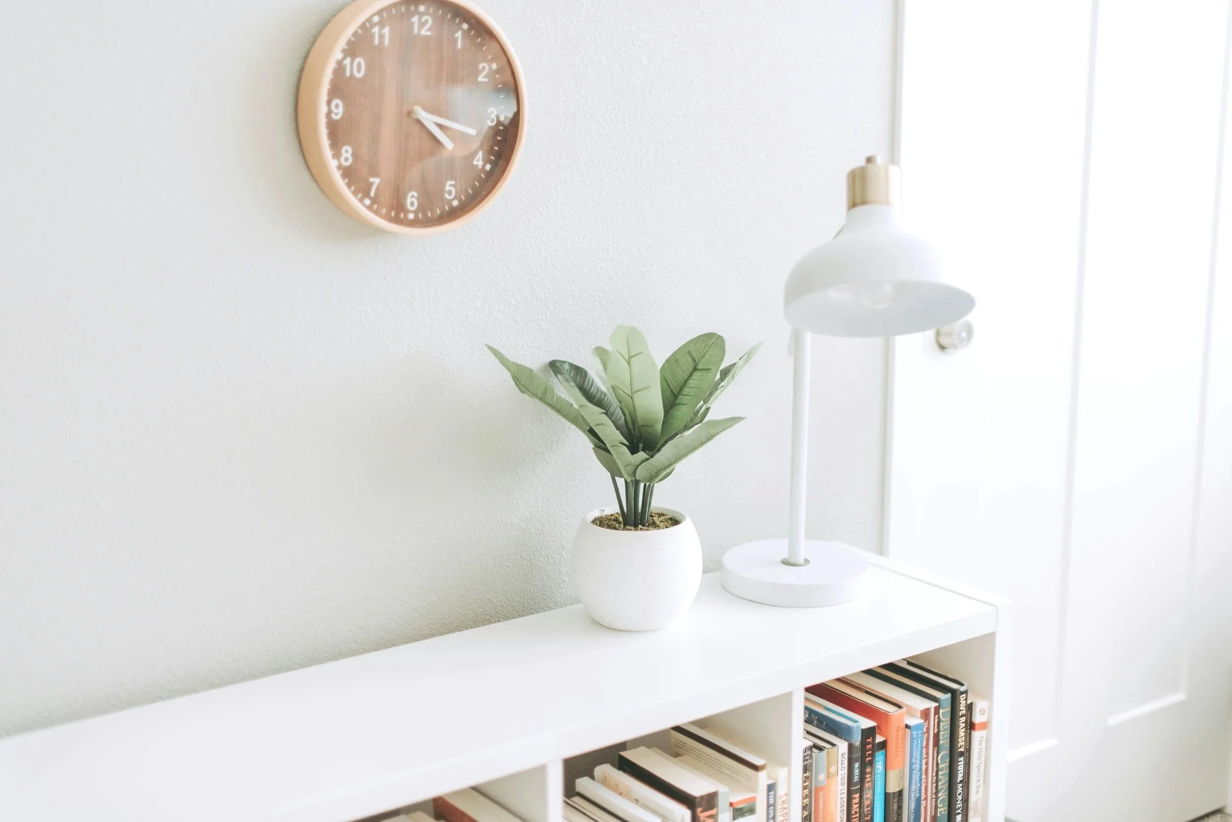 A white side table with a potted green plant, a white lamp, and a shelf with books underneath. On the wall above, there is a round wooden clock showing approximately 3:23.