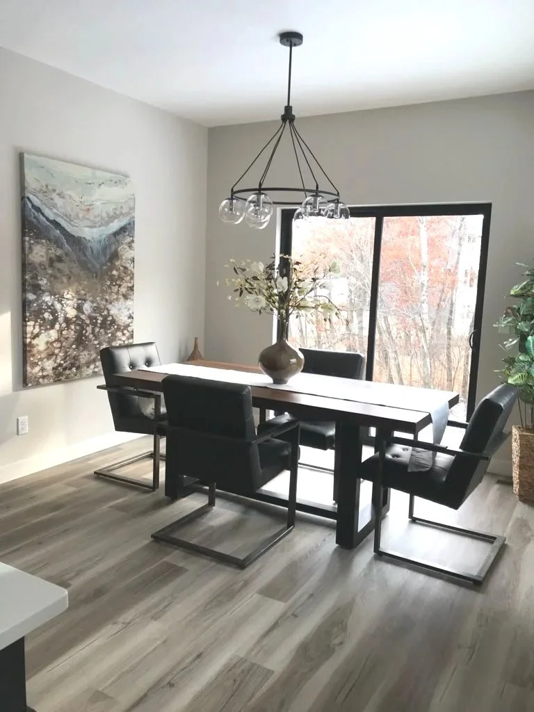 Modern dining room with a black wooden table, six black upholstered chairs, a large window with trees outside, a contemporary chandelier, a vase with flowers on the table, a large abstract painting on the wall, and a potted plant in a woven basket.