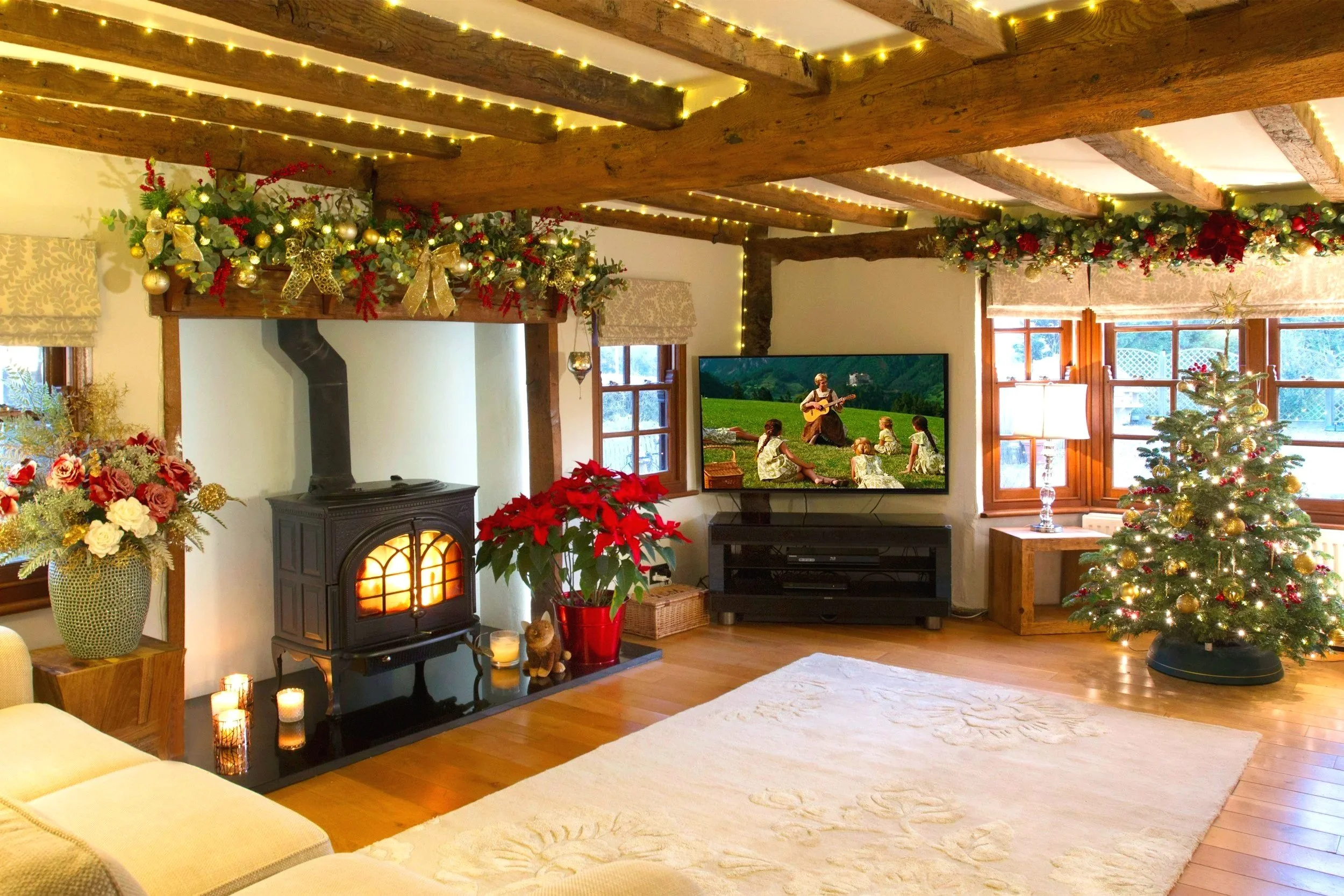 Living room decorated for Christmas with garlands and ornaments, a decorated Christmas tree, a fireplace with candles, and a television showing a scene of children listening to a woman playing guitar outdoors.