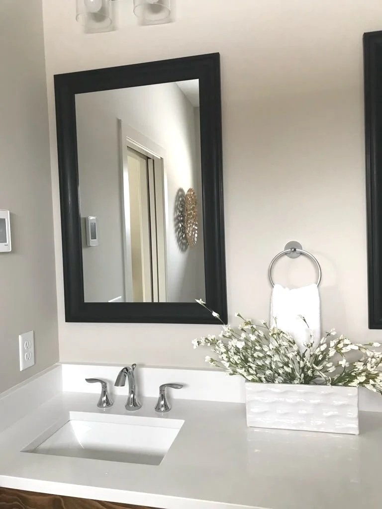 Bathroom vanity with a white countertop, sink, and silver faucet, a large black-framed mirror, a floral arrangement in a white pot, a white towel on a towel ring, and a wall-mounted thermostat.