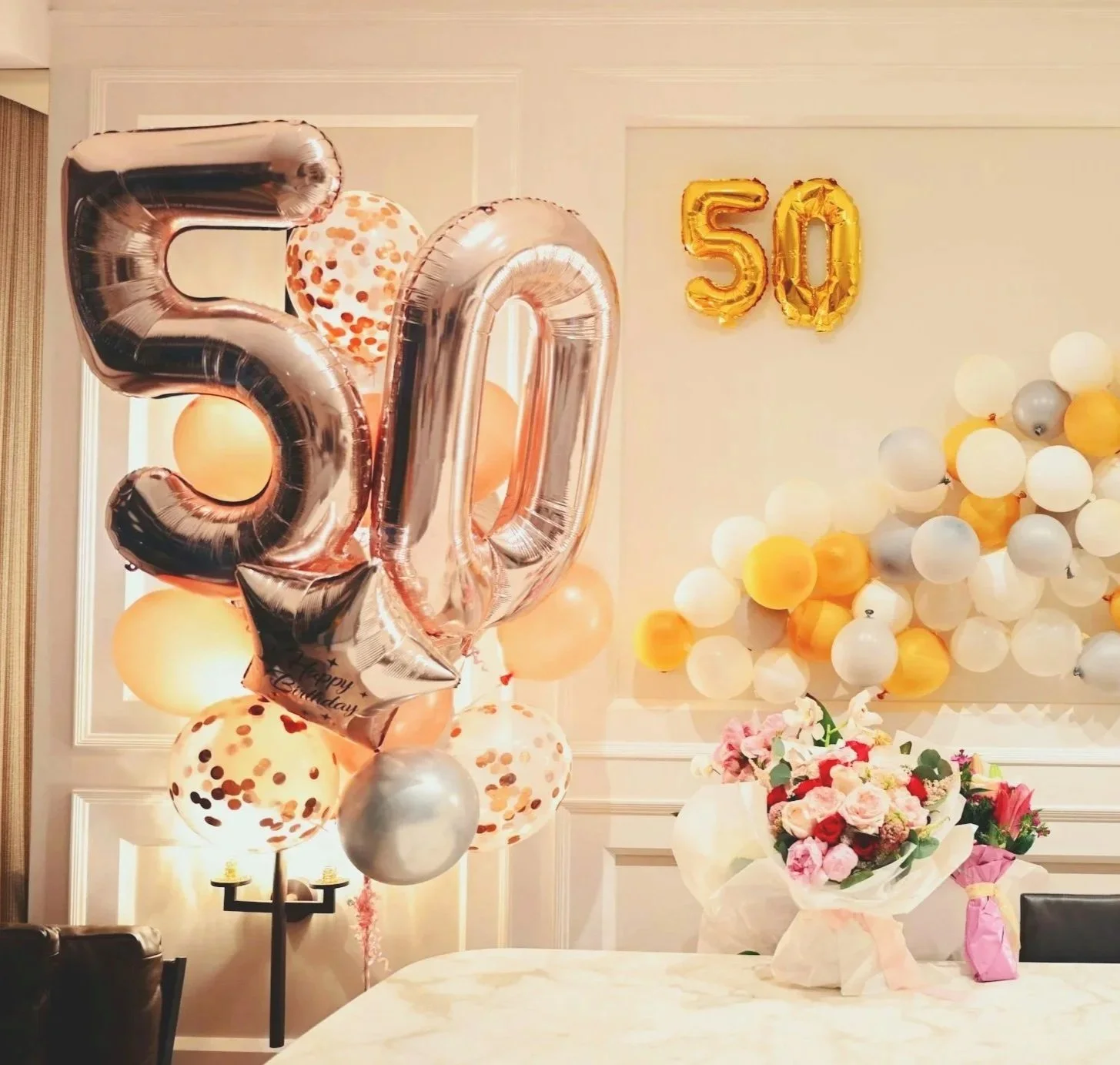Celebration decorations including large metallic balloons forming the numbers 50, surrounded by smaller balloons in gold, white, and silver, along with a bouquet of pink, red, and white flowers on a table.
