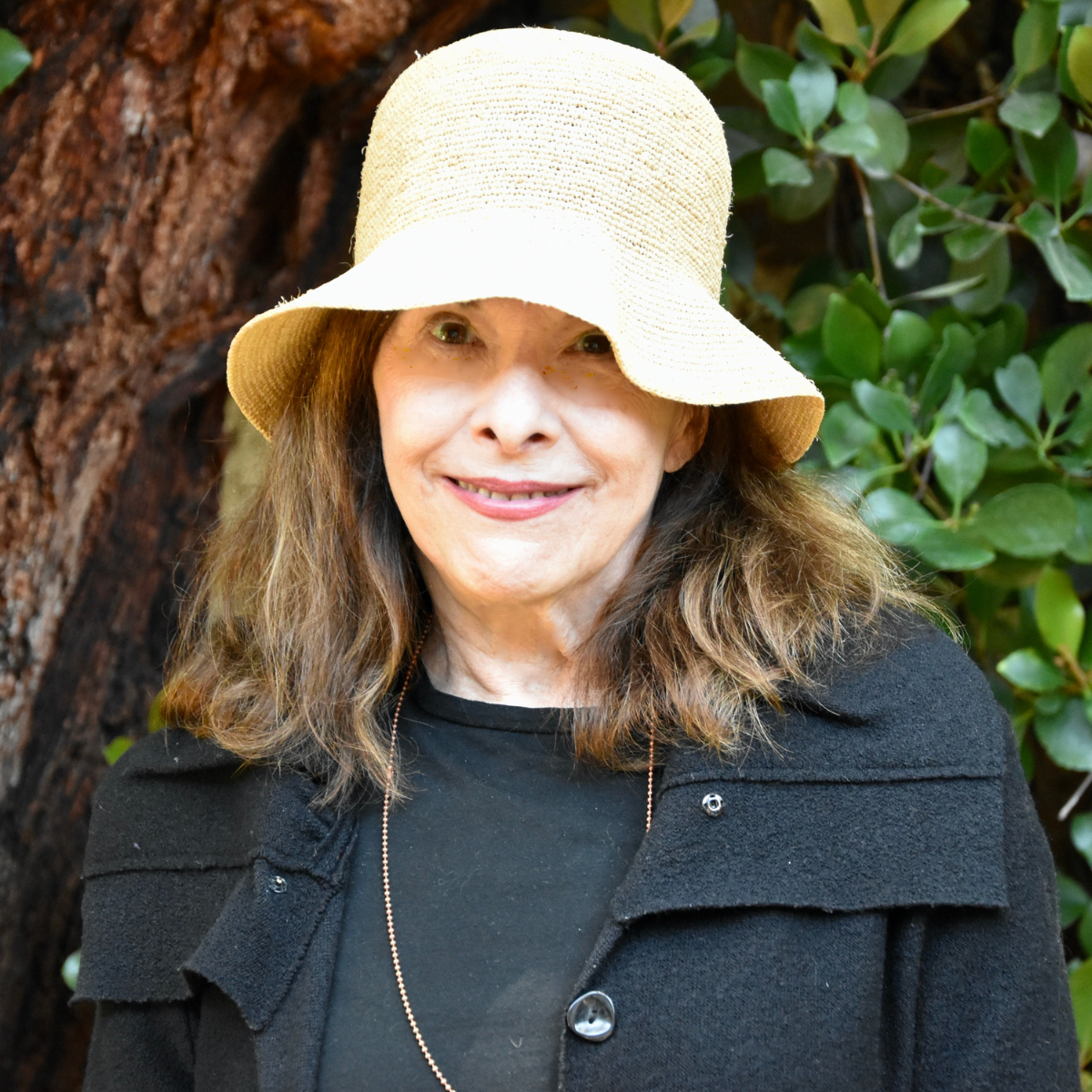 A woman wearing a beige sunhat and black coat, smiling outdoors with green foliage in the background.