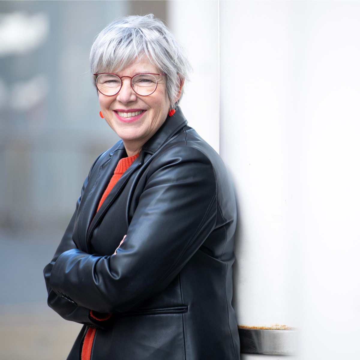 Smiling elderly woman with short gray hair and glasses, wearing a black leather jacket with orange shirt, leaning against a white wall.