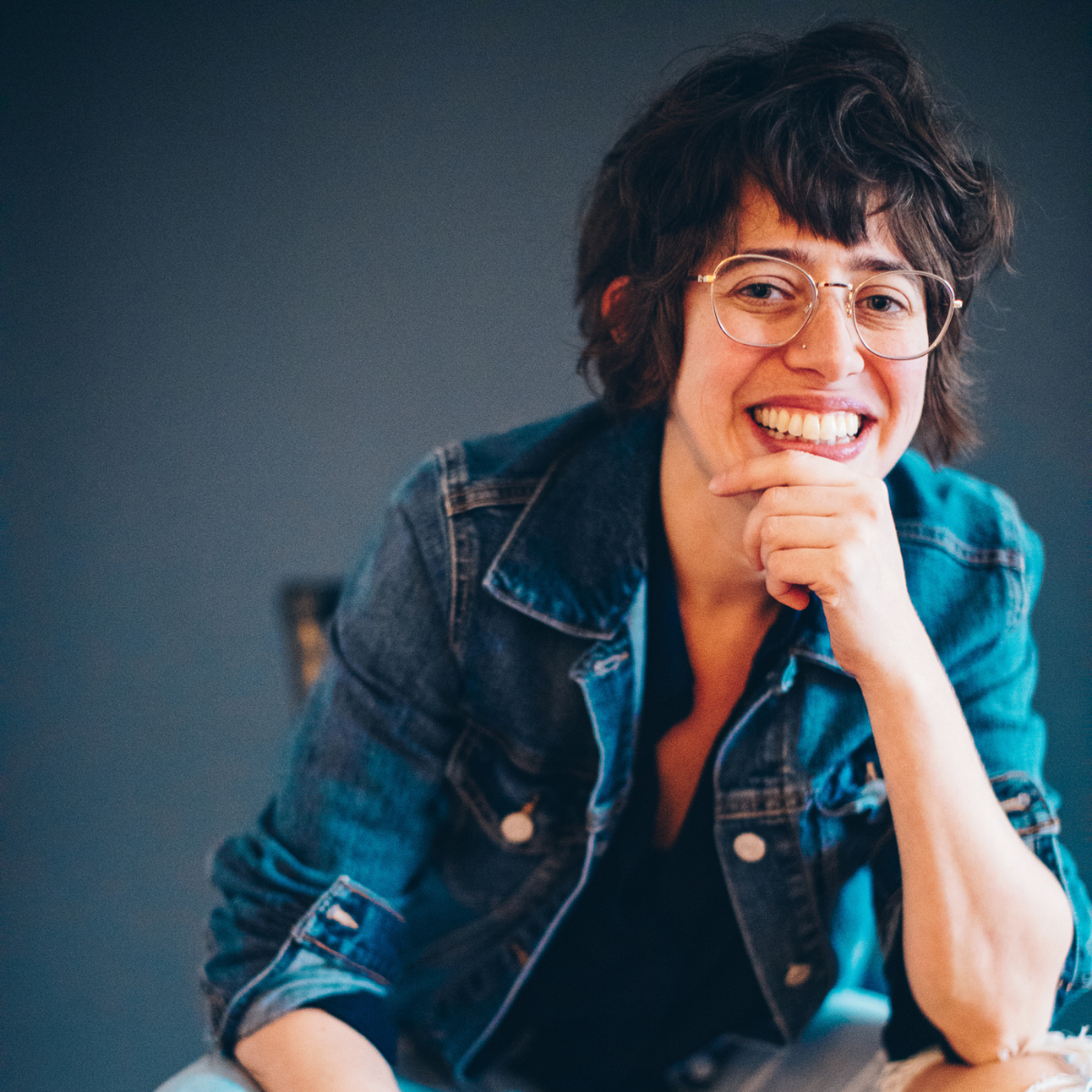 A young woman with glasses and short, curly hair smiling and sitting with her chin resting on her hand, wearing a denim jacket.