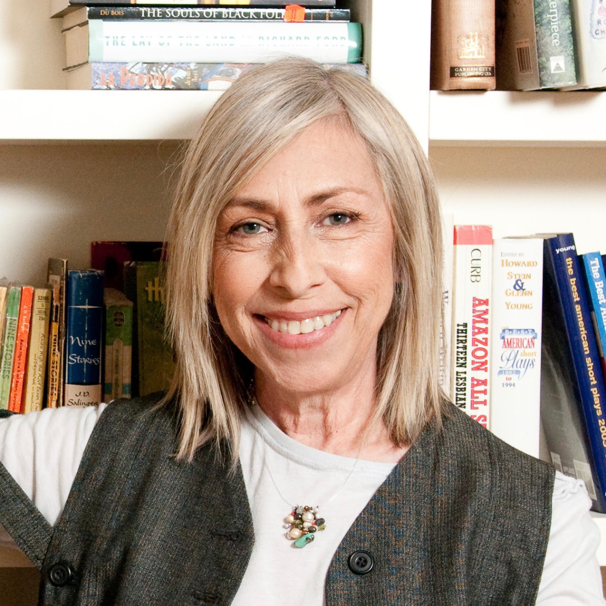 A woman with shoulder-length gray hair smiling, wearing a white shirt, a gray vest, and a colorful jewelry necklace, standing in front of a bookshelf filled with various books.