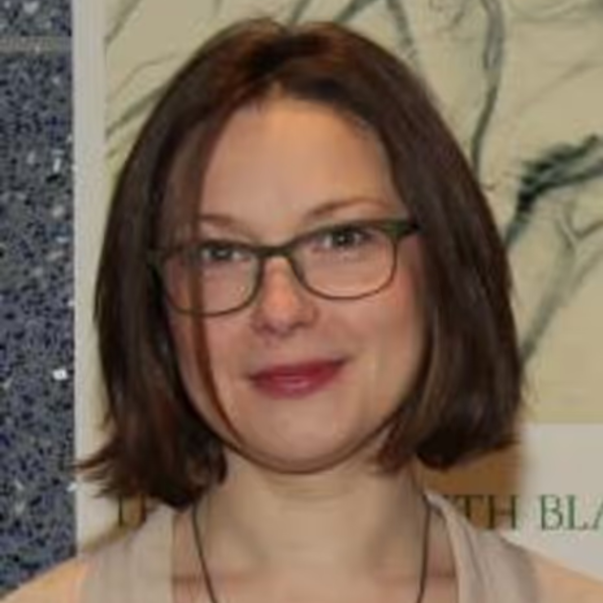 A woman with shoulder-length brown hair, glasses, and a light-colored top, smiling in front of a background with abstract artwork and text.