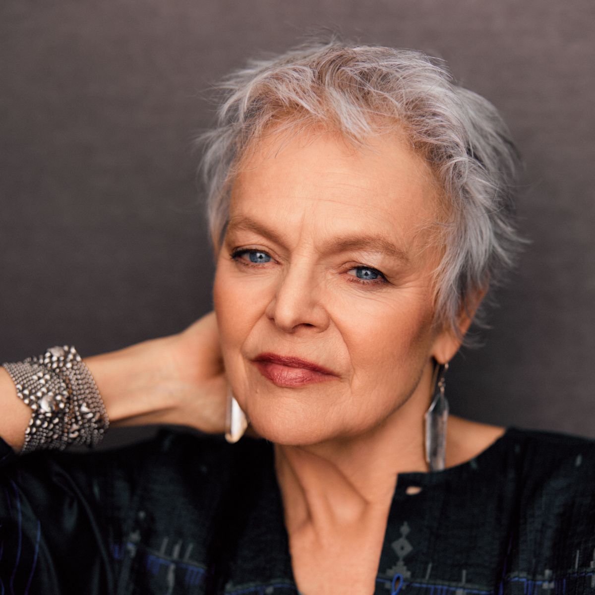 Close-up portrait of an older woman with short, gray hair, wearing makeup, a black top, and large earrings. She has a confident expression and is touching her neck with her right hand, which is adorned with a beaded bracelet.