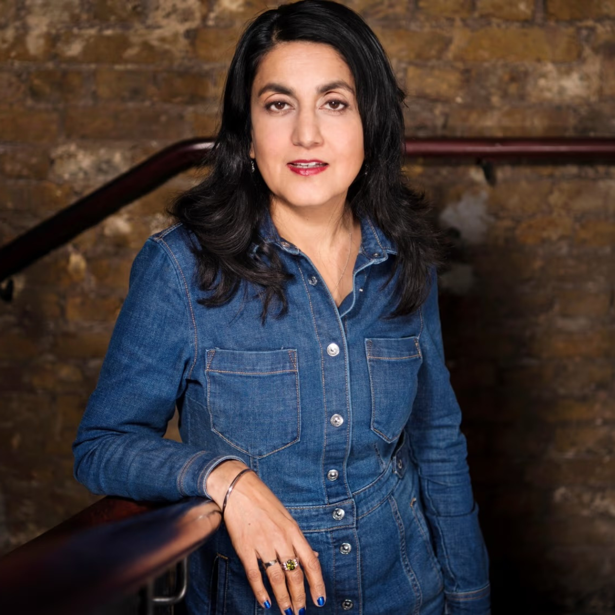 A woman with black hair wearing a denim dress standing in front of a brick wall background, leaning on a wooden handrail.