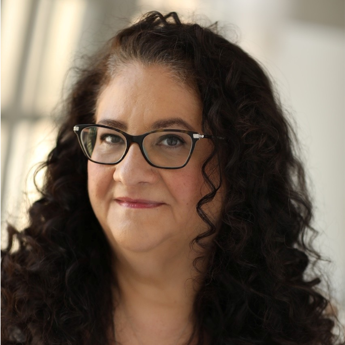 Close-up of a woman with curly dark hair, wearing glasses, smiling slightly, indoors near natural light.