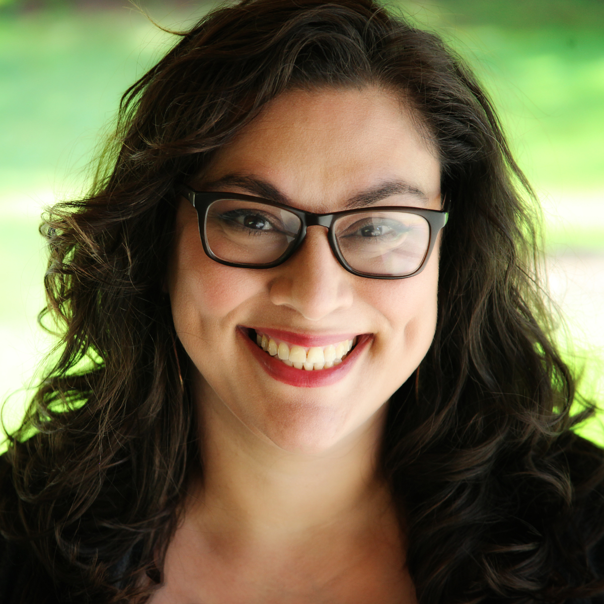 A woman with long wavy dark hair and dark-rimmed glasses smiles at the camera. She wears a dark top open at the neck. Blurred, light-filled folliage and grass in the background.