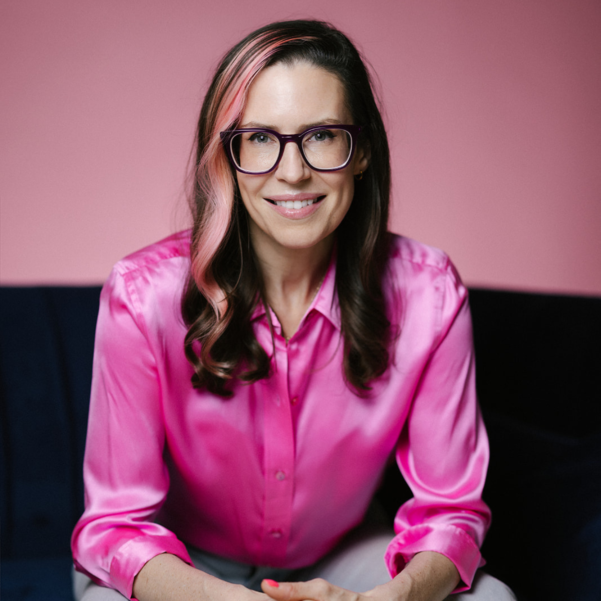 A woman with long dark hair and pink highlights, wearing glasses and a shiny pink blouse, sitting on a dark blue couch with a pink background.