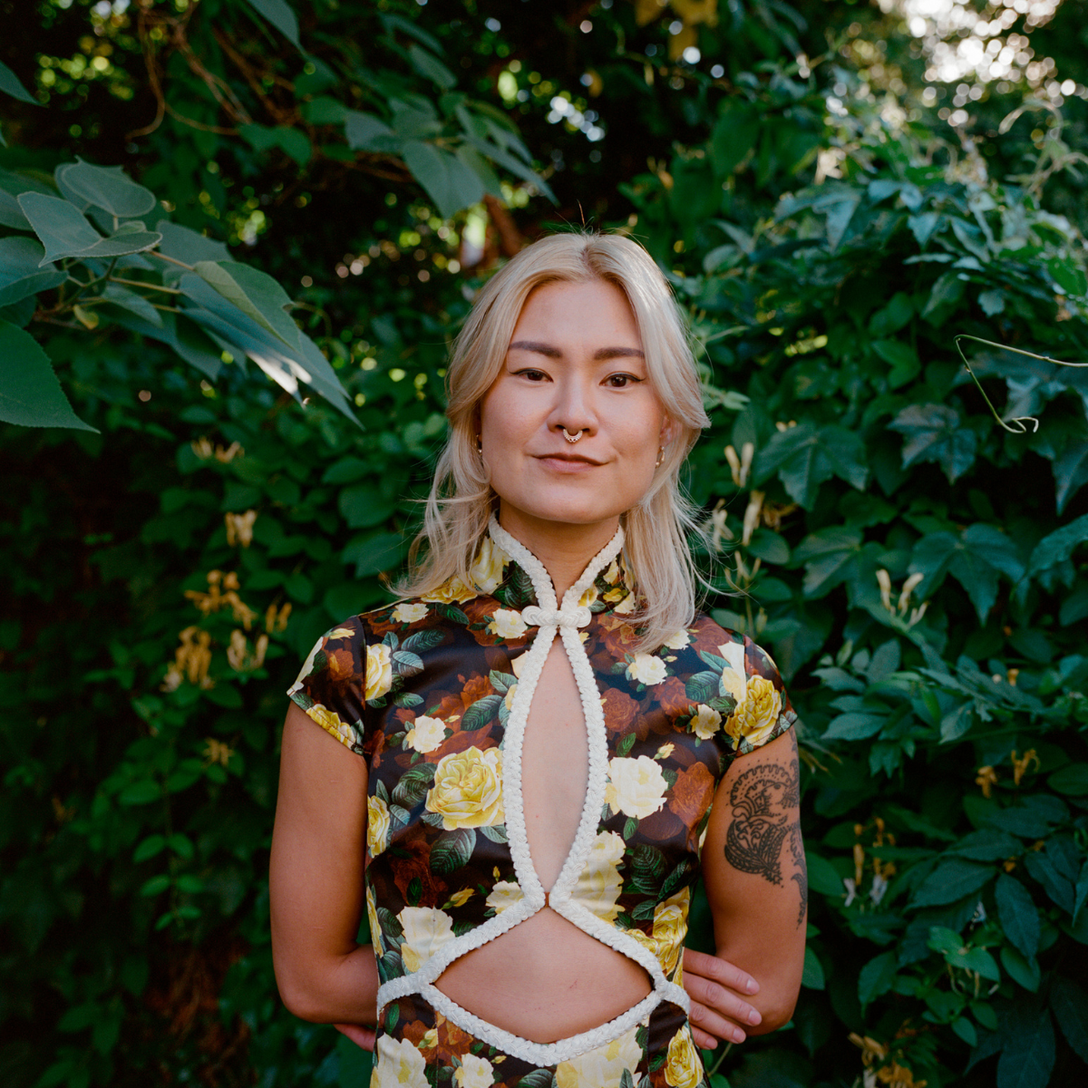 A woman with blonde hair and a septum piercing standing in front of lush green foliage. She is wearing a floral top with cutouts and a decorative white trim.