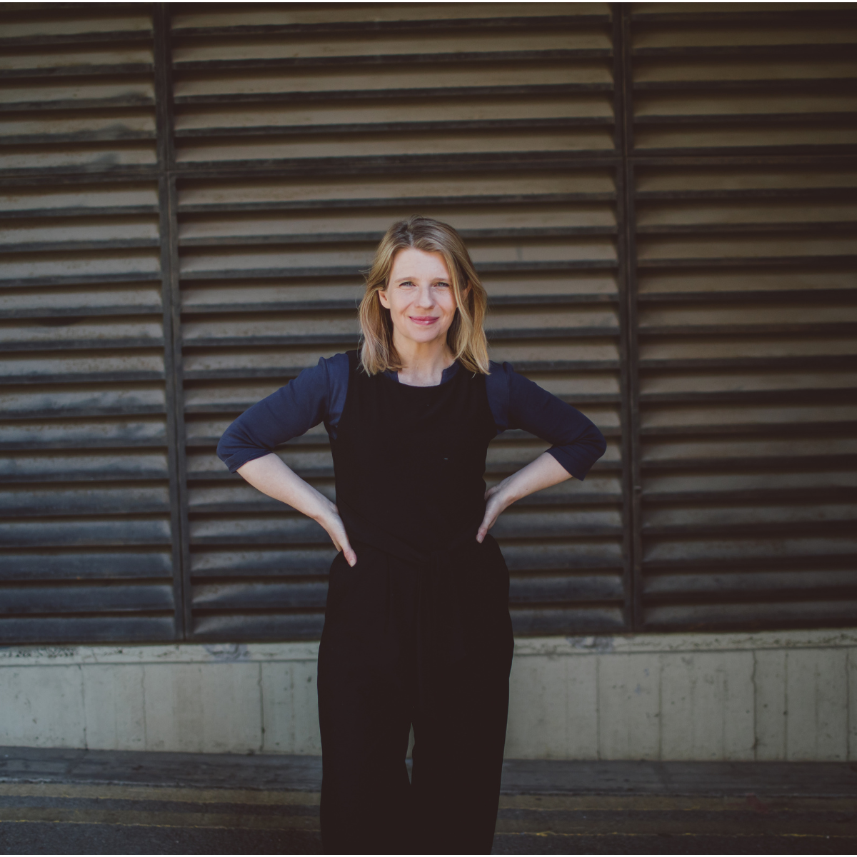 A woman with shoulder-length blonde hair, wearing a black top with rolled-up sleeves, standing with her hands on her hips in front of wooden louvered panels.