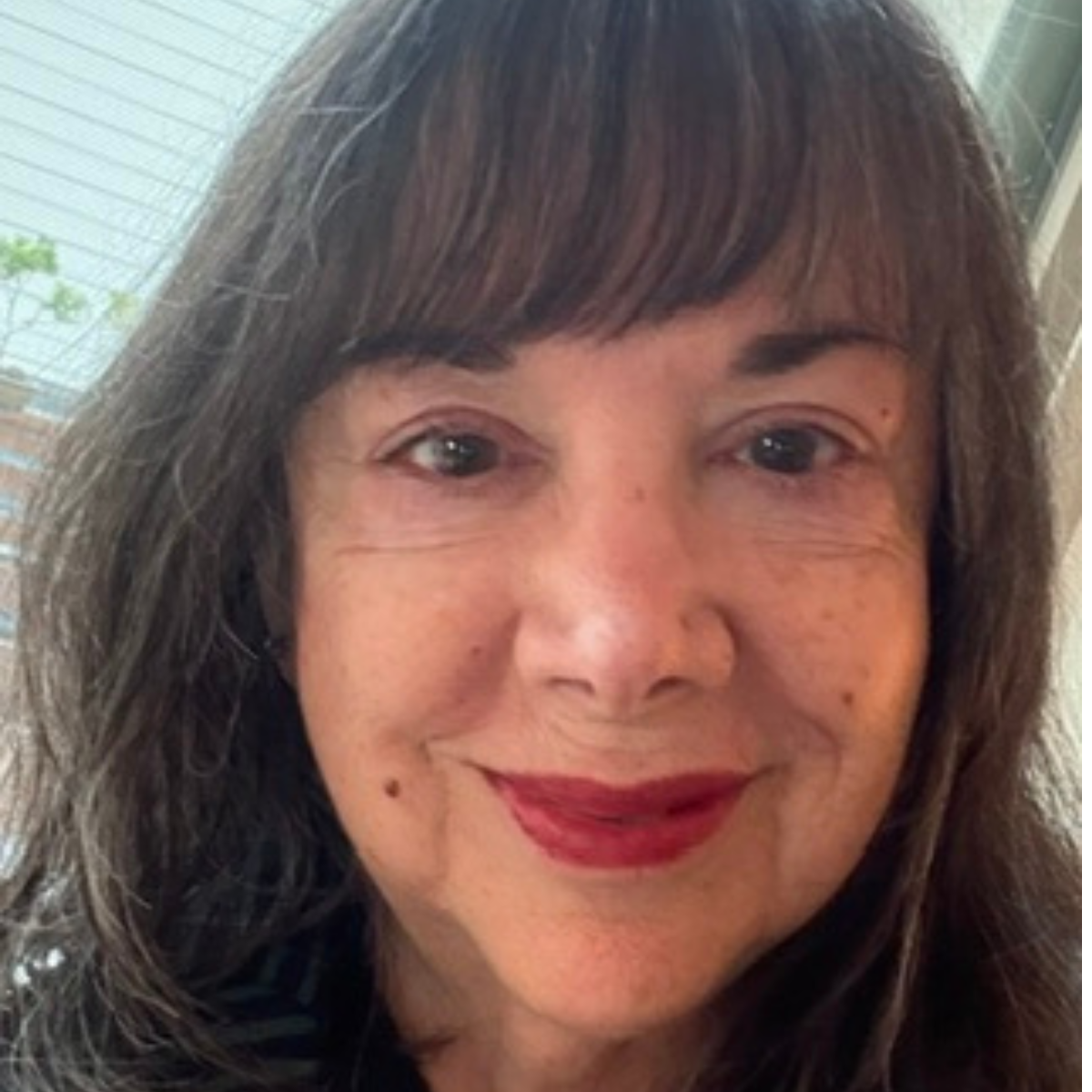 Close-up of a smiling woman with shoulder-length brown hair and red lipstick, indoors near a window with blinds and some greenery visible outside.