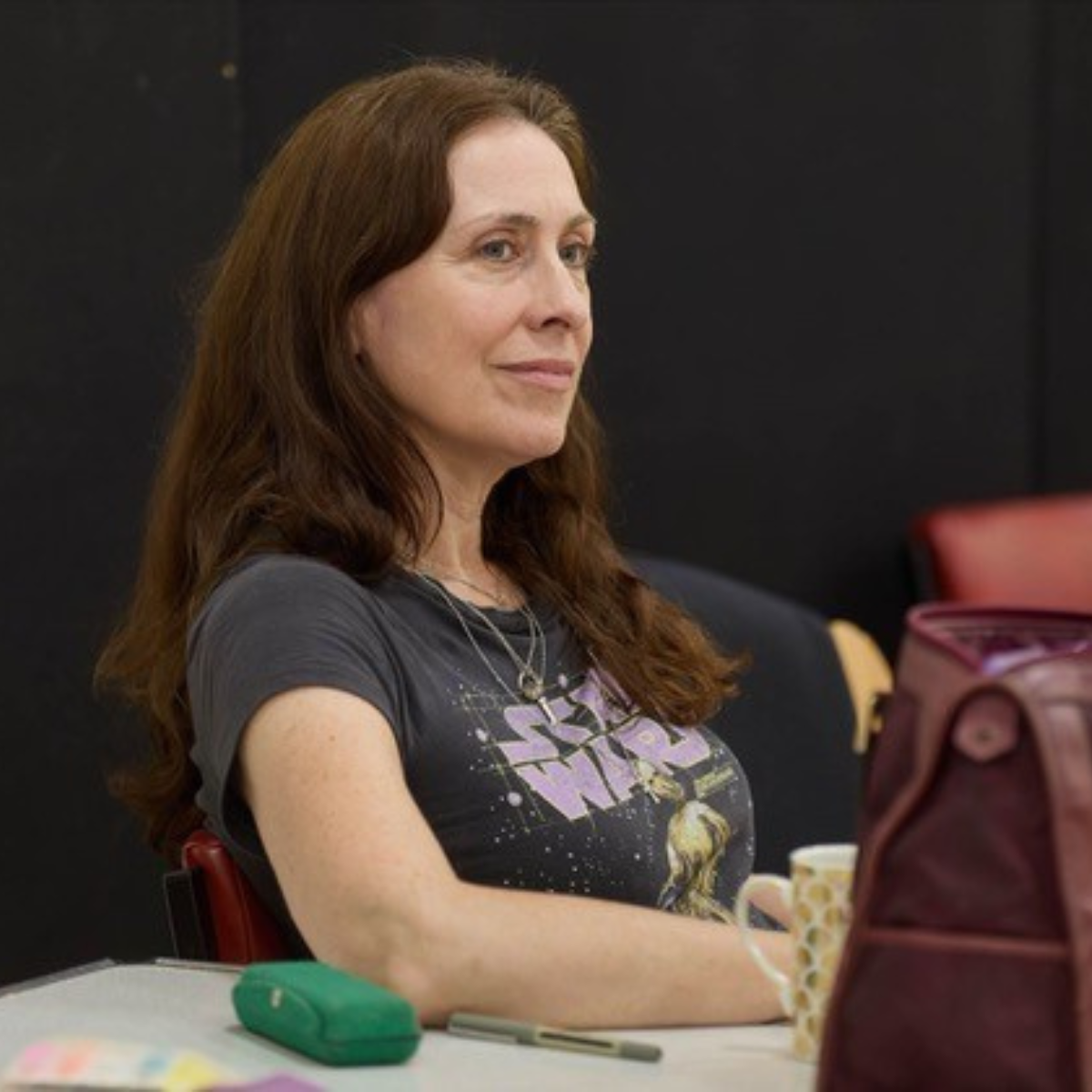 A woman with long, wavy reddish-brown hair sitting at a table, wearing a grey t-shirt with a graphic design, gold necklaces, holding a cup with a yellow polka dot pattern, in a room with black walls and red chairs.