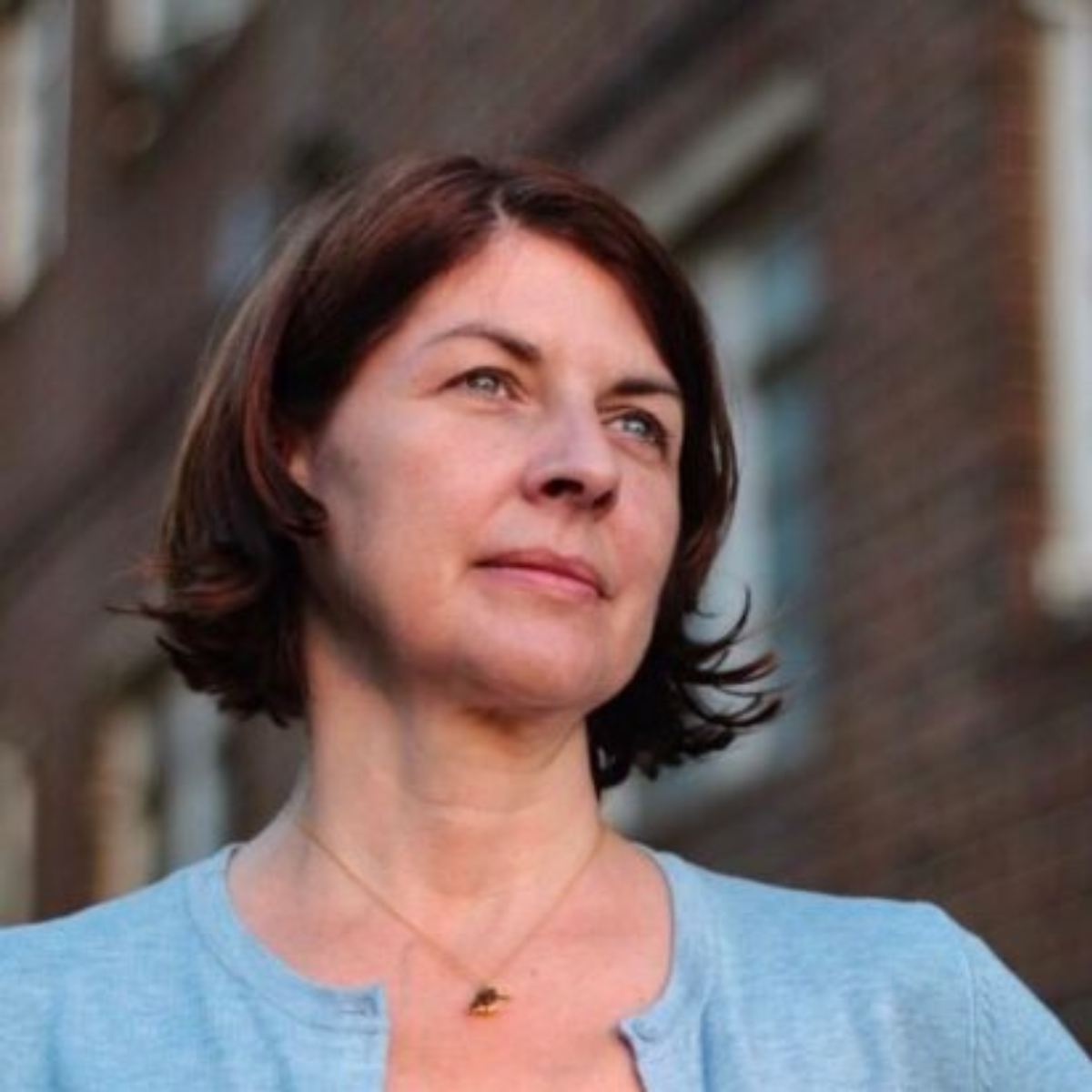 A middle-aged woman with short brown hair, wearing a light blue top and a small gold necklace, standing outdoors in front of a brick building and looking into the distance.
