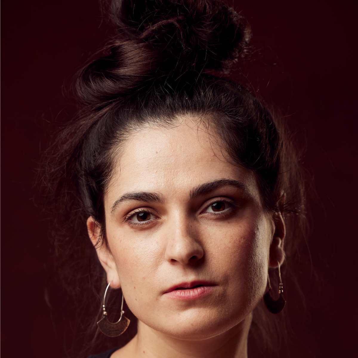 Close-up of a young woman with dark hair styled in a messy bun, wearing earrings, with a serious expression against a dark red background.