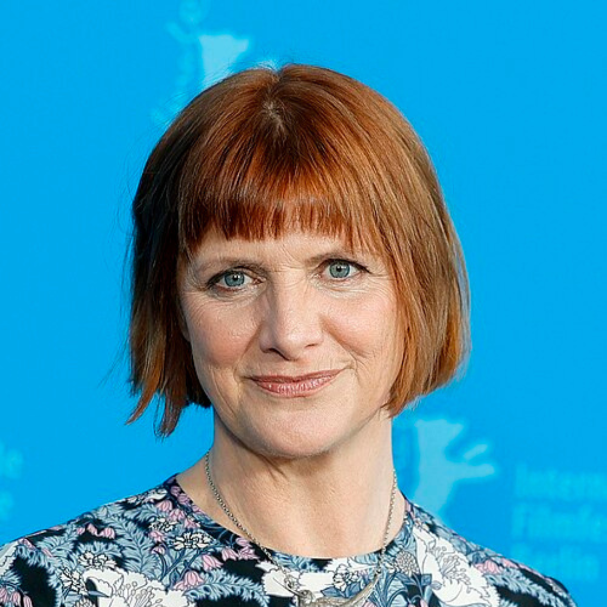 A woman with short red hair and blue eyes, wearing a floral patterned top and a silver necklace, standing in front of a blue background.
