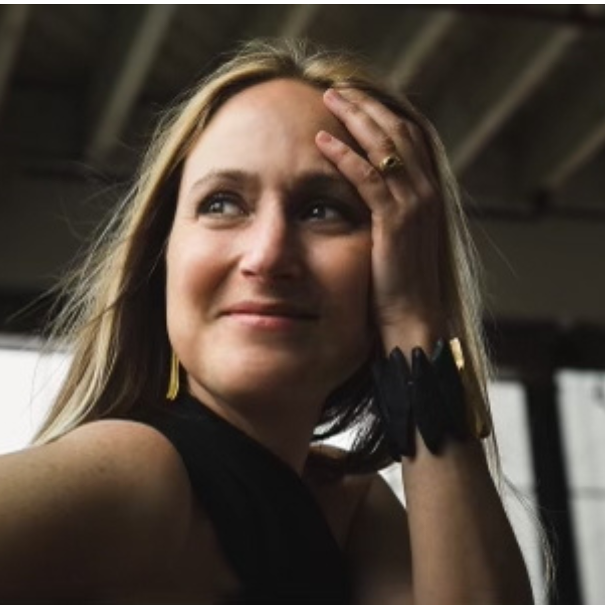 A woman with long blonde hair smiling and resting her hand on her forehead, wearing a black sleeveless top and jewelry, indoors with a blurred ceiling background.