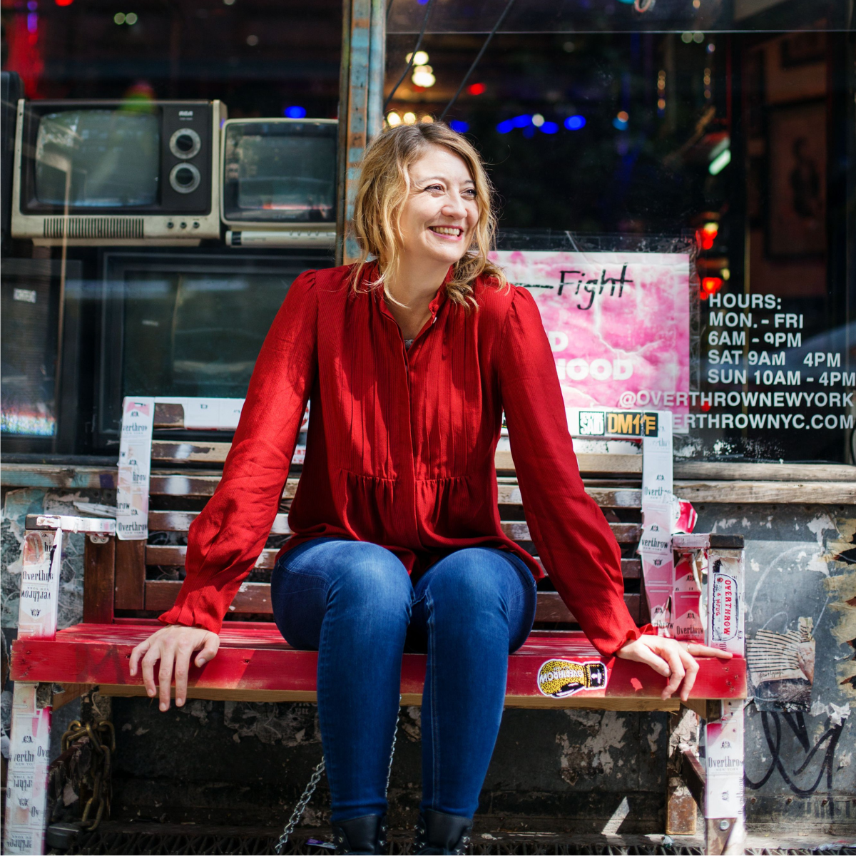 A woman with blonde hair wearing a red blouse and blue jeans, sitting on a worn wooden bench outside a storefront with neon signs and graffiti, smiling and looking to her right.