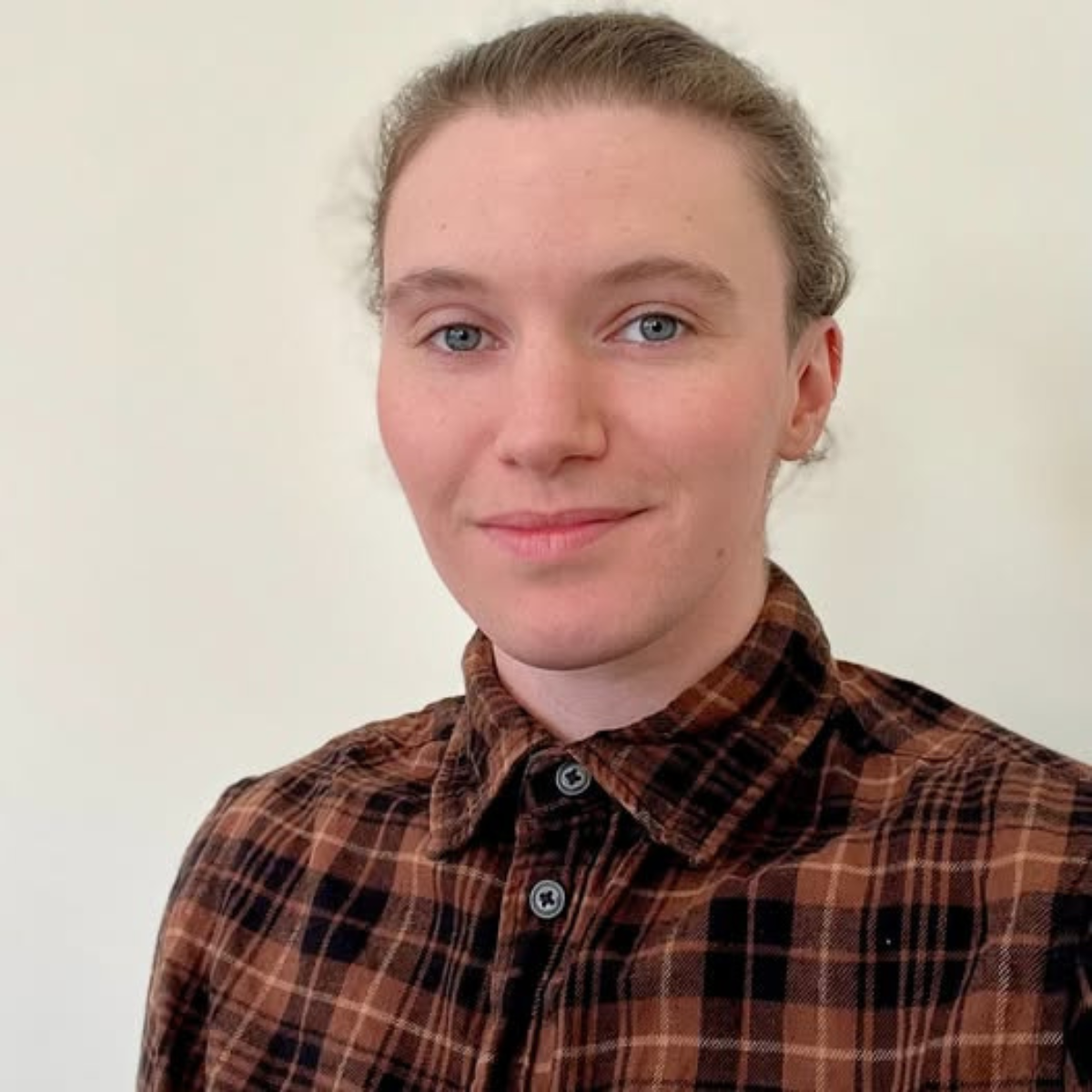 Portrait of a young woman with light skin and blue eyes, wearing a brown plaid shirt, standing against a plain off-white wall.