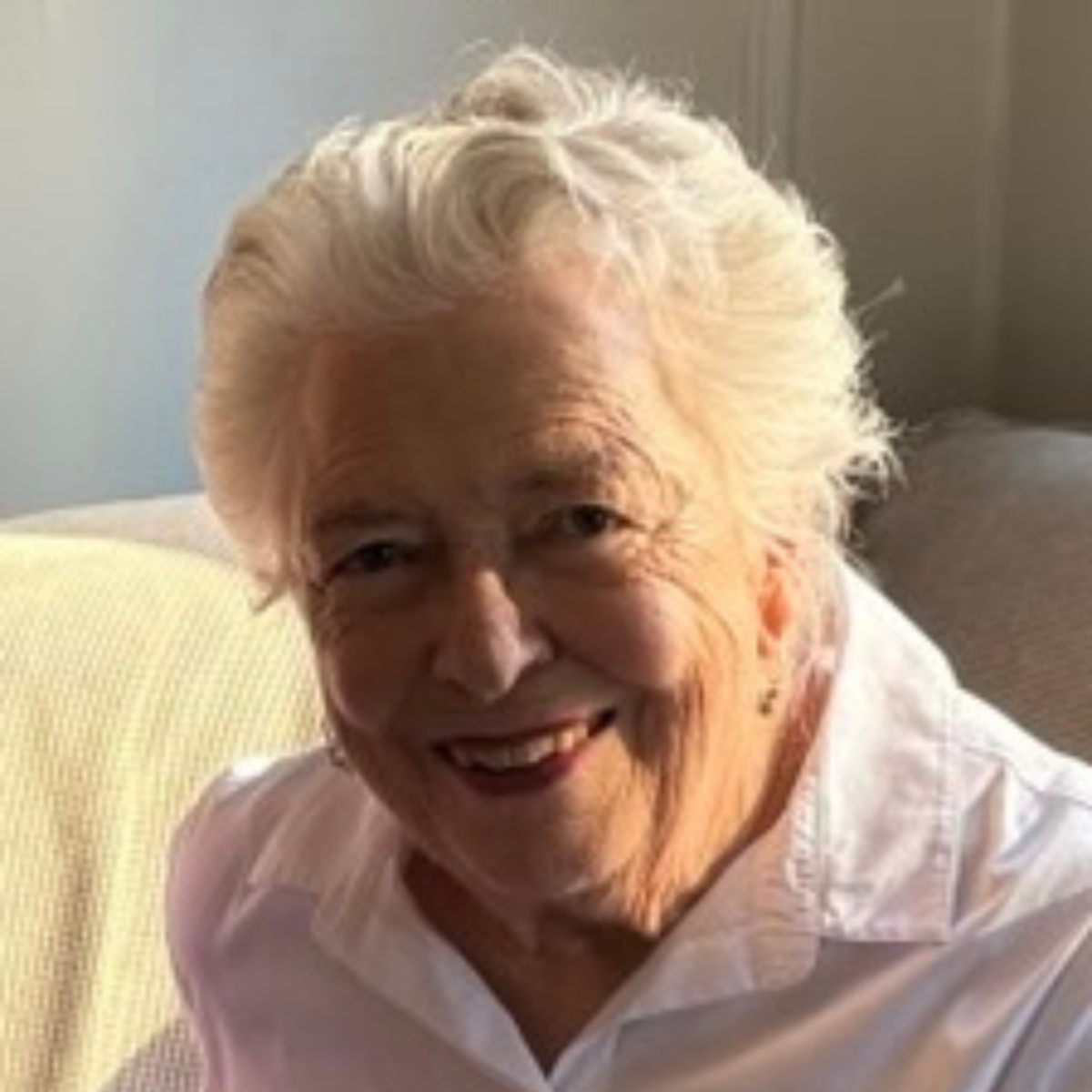 An elderly woman with white hair smiling and wearing a white shirt, sitting on a light yellow and gray couch.