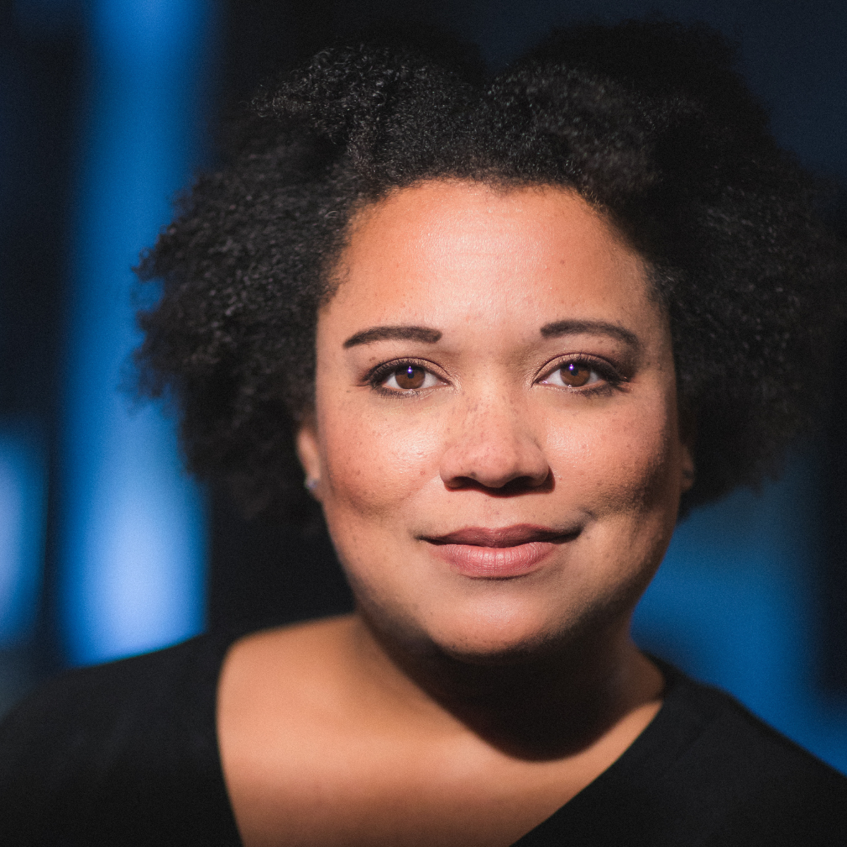 A Black woman with short curly dark hair looks at the camera, smiling lightly. She wears a dark top and there is a dark background with blurred blue light.