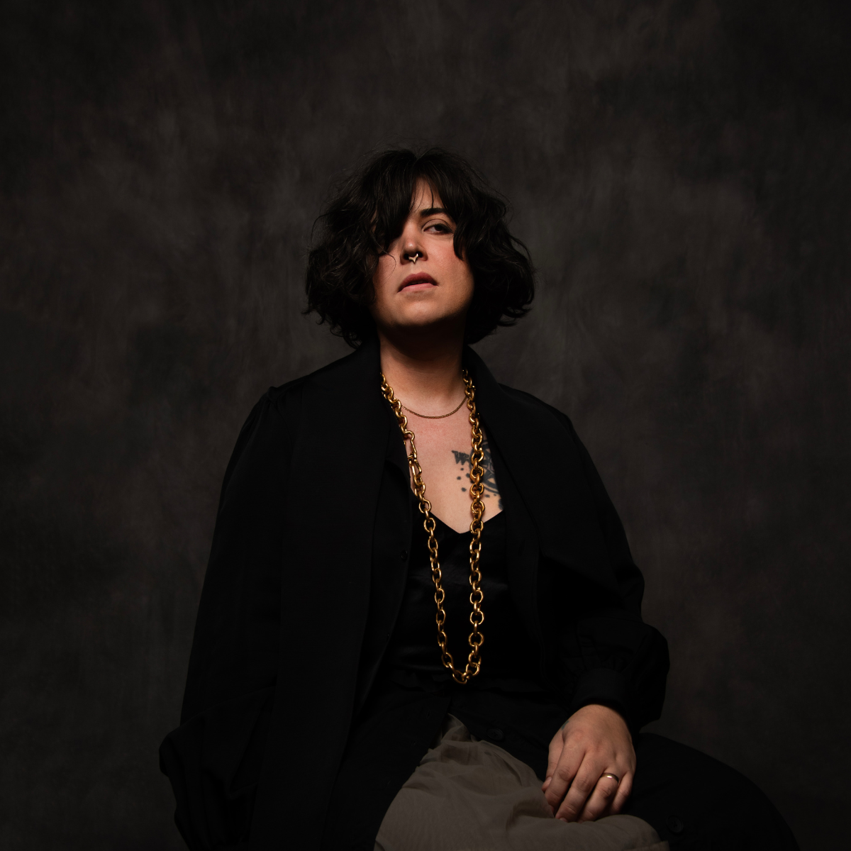 A person with shoulder-length dark curly hair, wearing a black shirt and jacket, accessorized with gold chains and a septum piercing, sitting against a dark textured background.