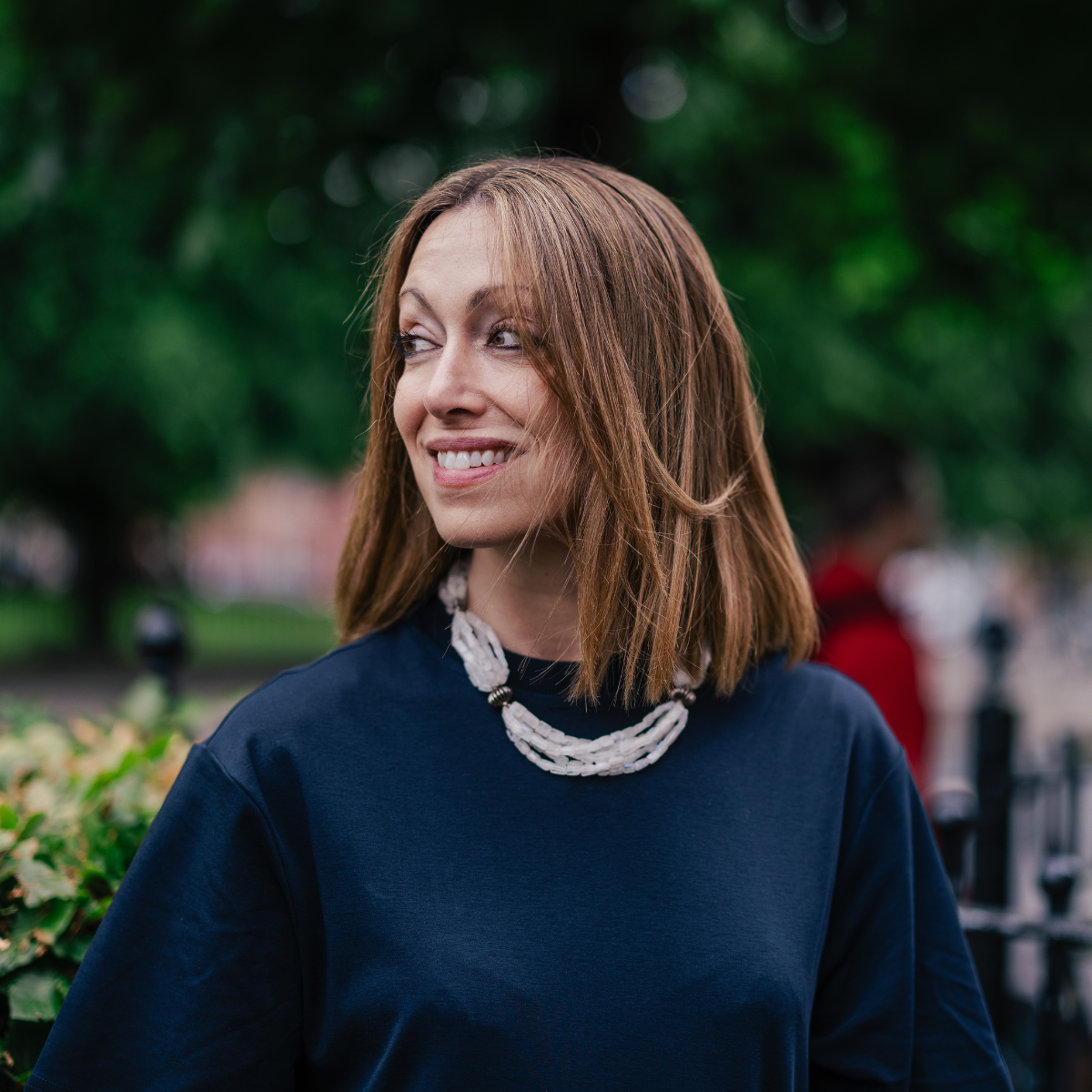 A woman with shoulder-length auburn hair is smiling and looking to her left outdoors, wearing a dark blue top and a white beaded necklace. The background includes green trees and other people.