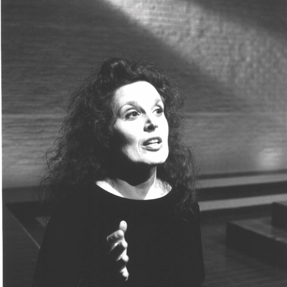 Black and white photo of a woman with curly hair speaking, her hand raised in a gesture, in a room with wooden walls.