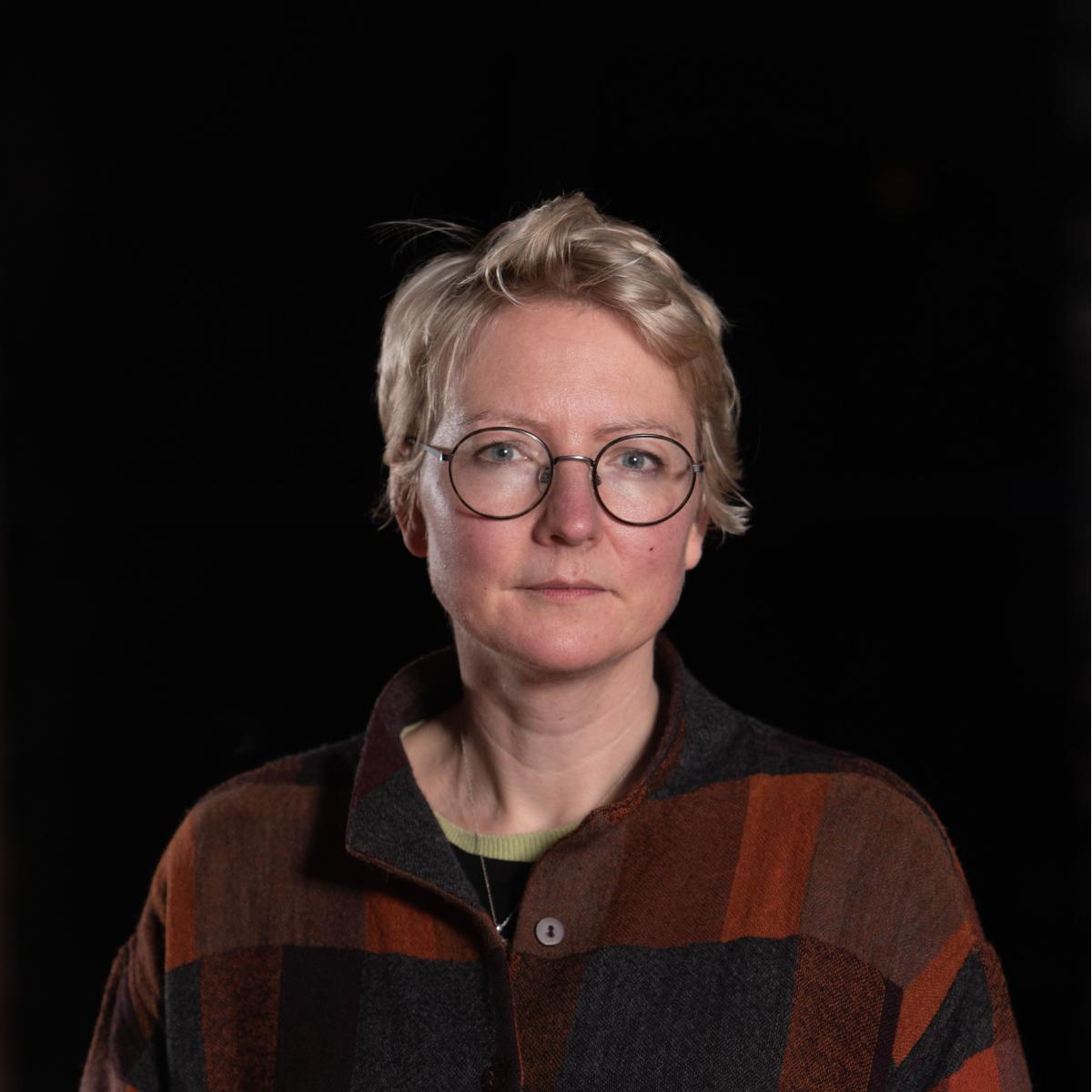 Portrait of a woman with short blond hair, round glasses, and a serious expression, wearing a checkered jacket in shades of brown and orange against a black background.