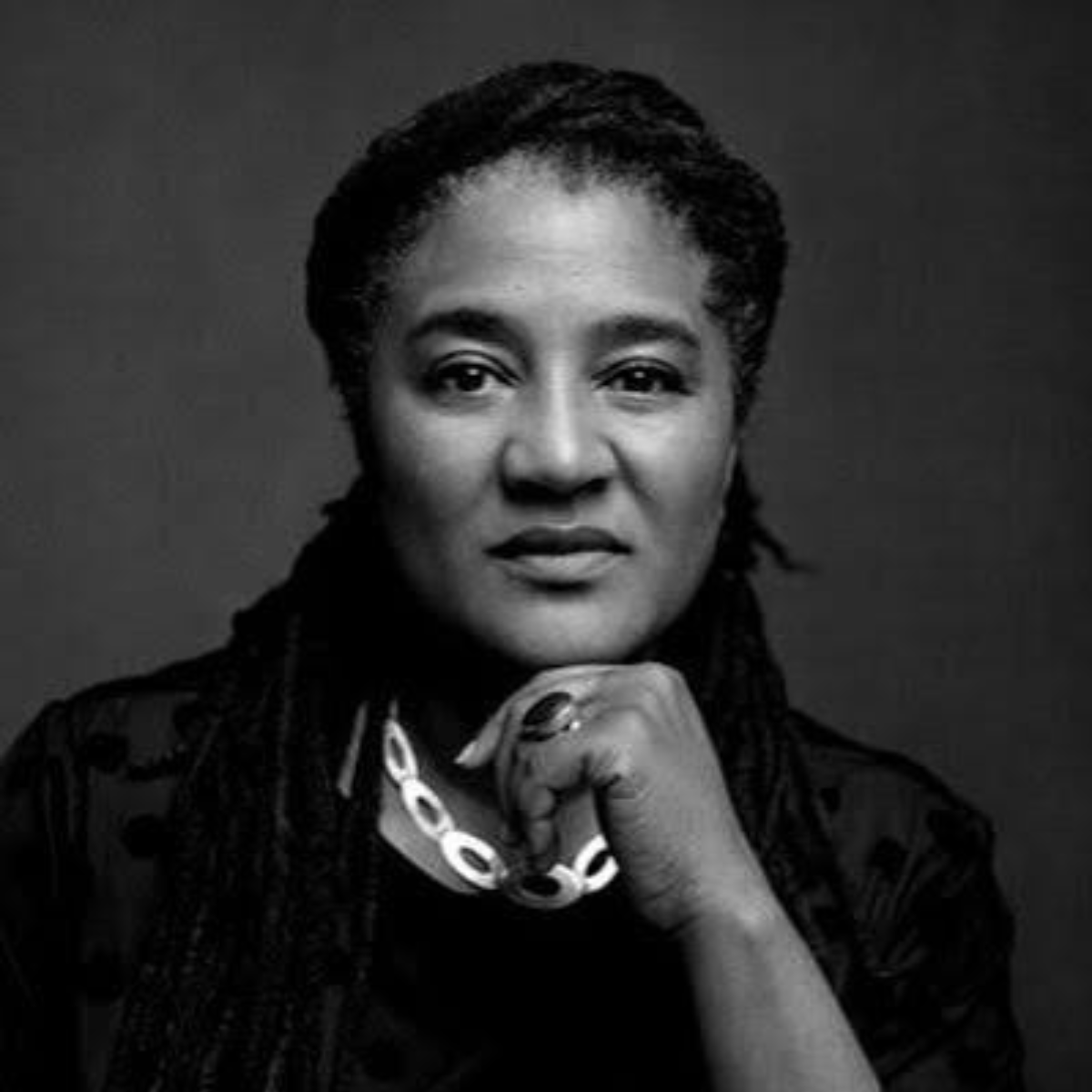Black and white portrait of a woman with braided hair, resting her chin on her hand, wearing a chunky necklace and ring, looking directly at the camera.