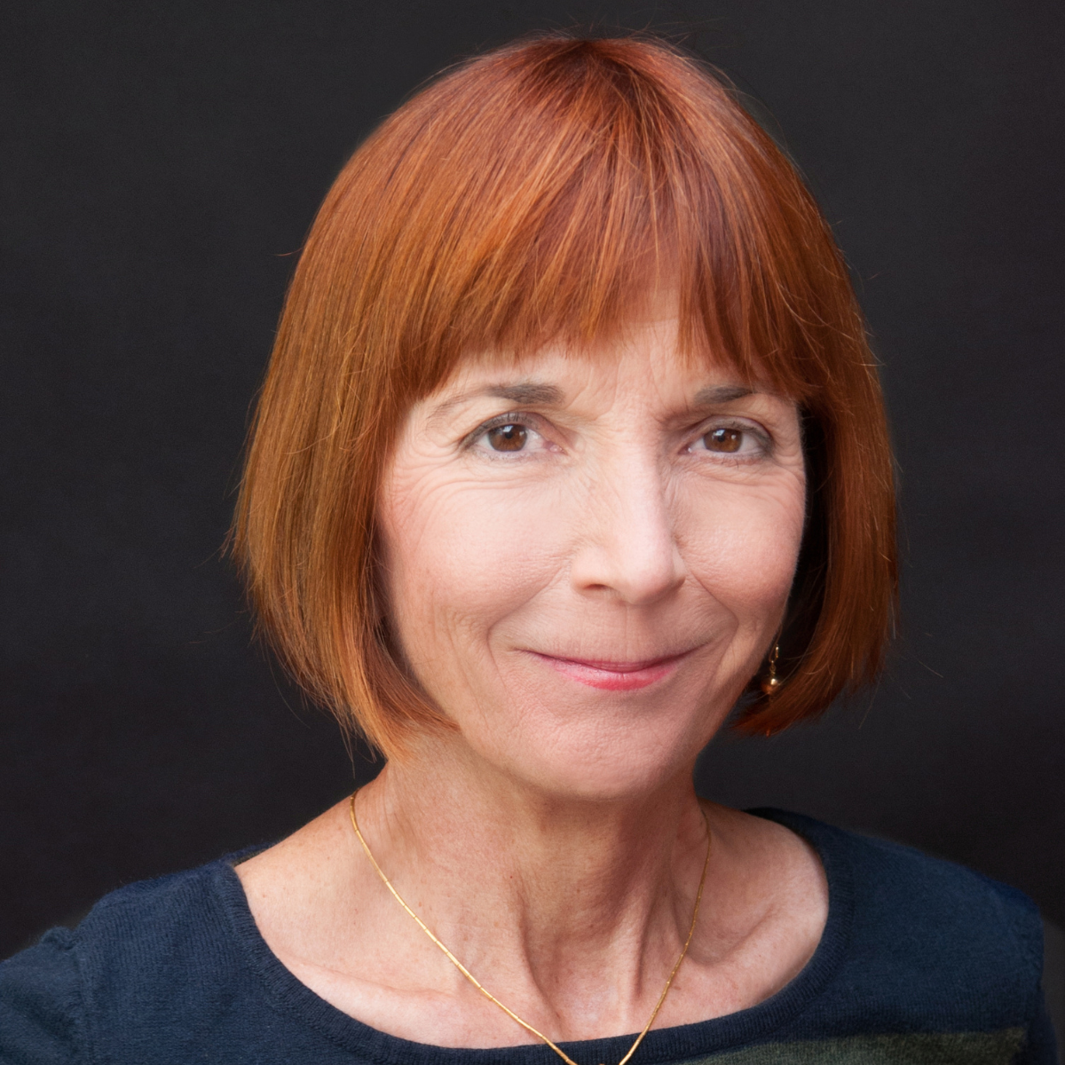 Close-up portrait of a middle-aged woman with short red hair, wearing a dark top, gold necklace, and earrings, smiling against a black background.
