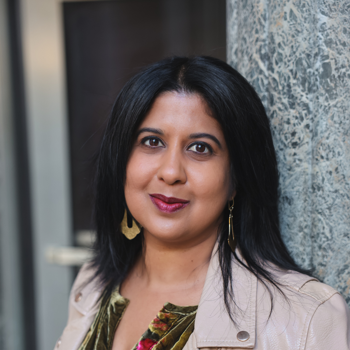 A woman with long, straight dark hair stands in 3/4 profile and looks at the camera with a light smile. She wears large dangling gold earrings, a green and cream floral top, and an off-white leather jacket. She stands against a stone building.