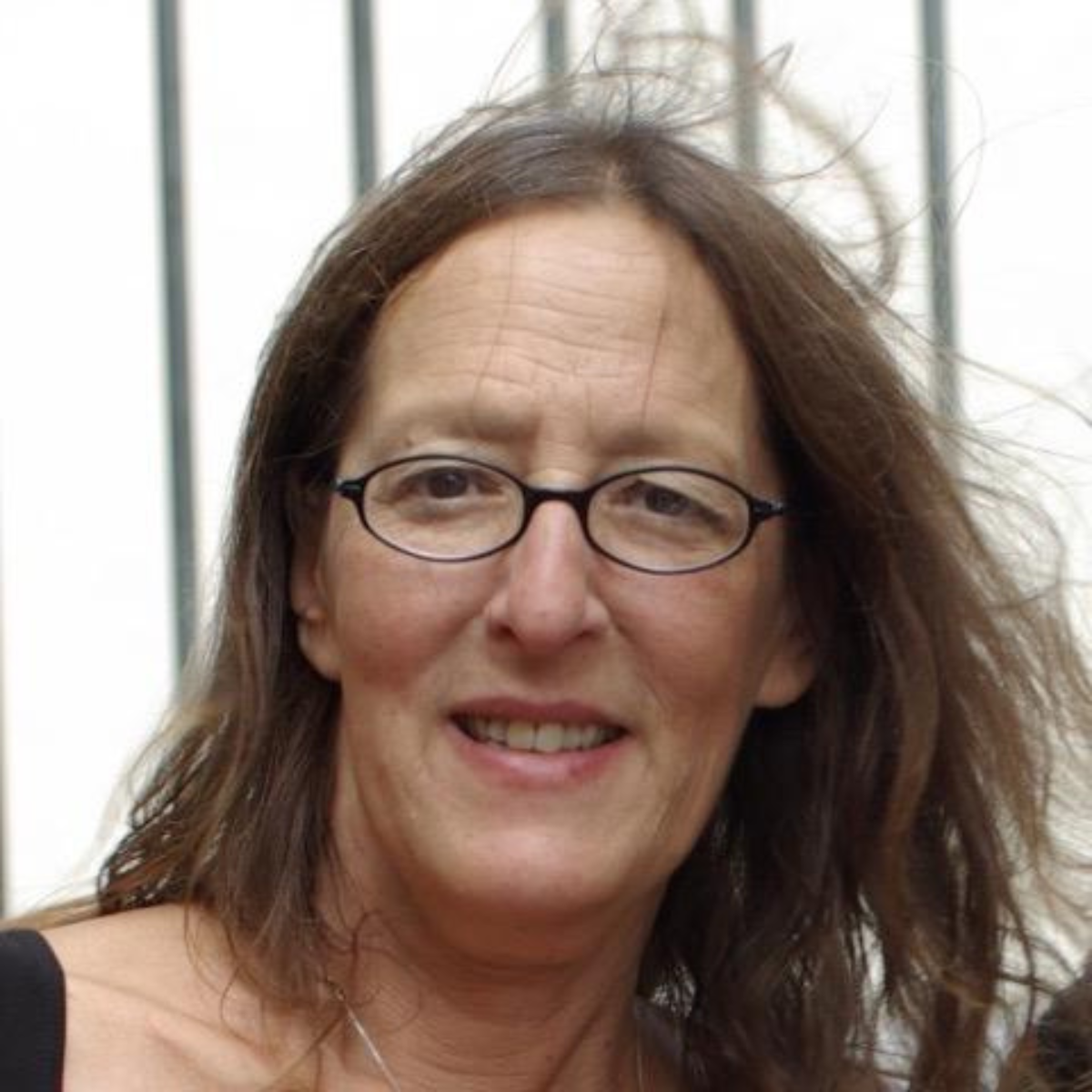A middle-aged woman with glasses and long, wavy brown hair smiling at the camera, with a blurred outdoor background.