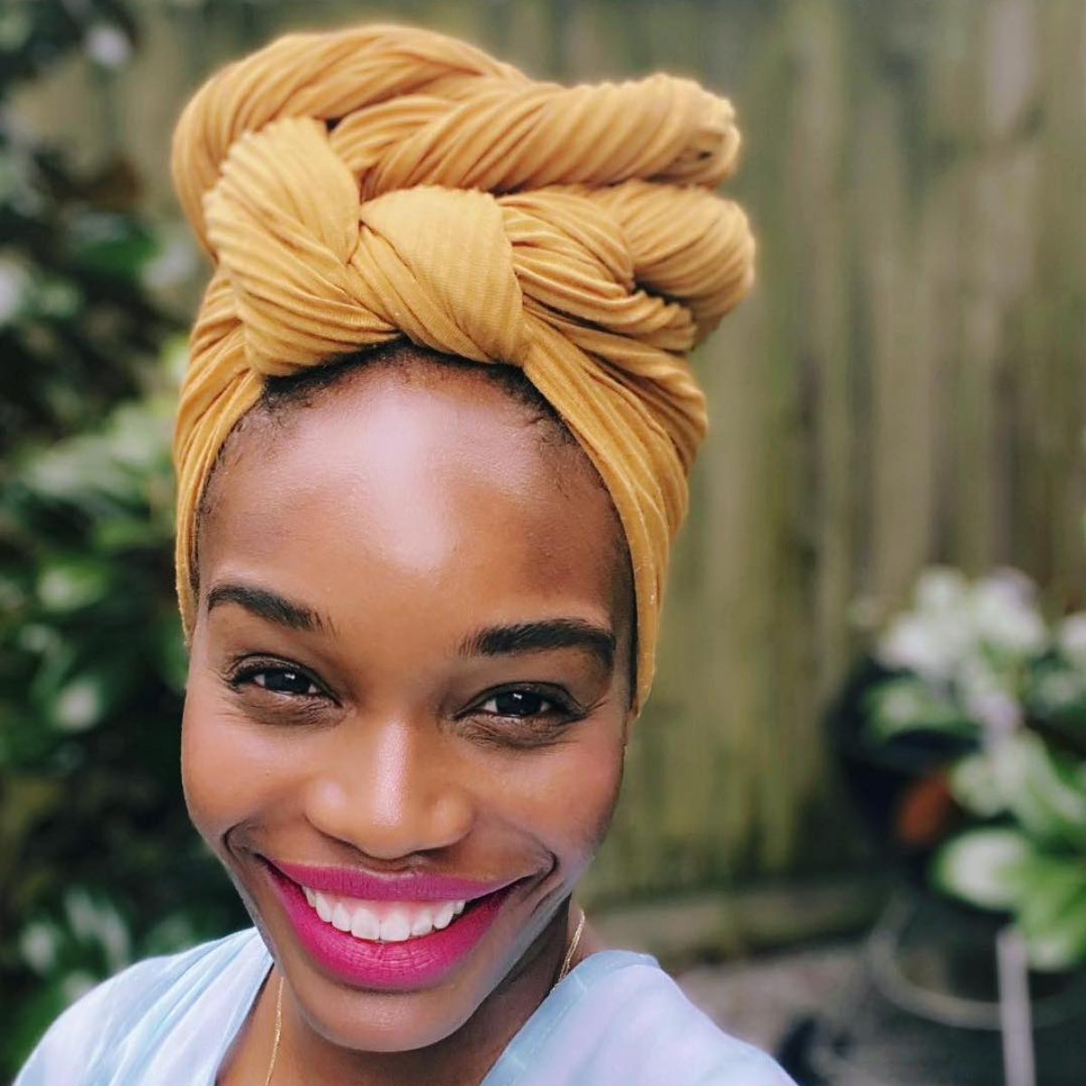 A woman smiling with bright pink lipstick wearing a mustard yellow headwrap and a light blue top. She is outdoors with a blurred garden background.