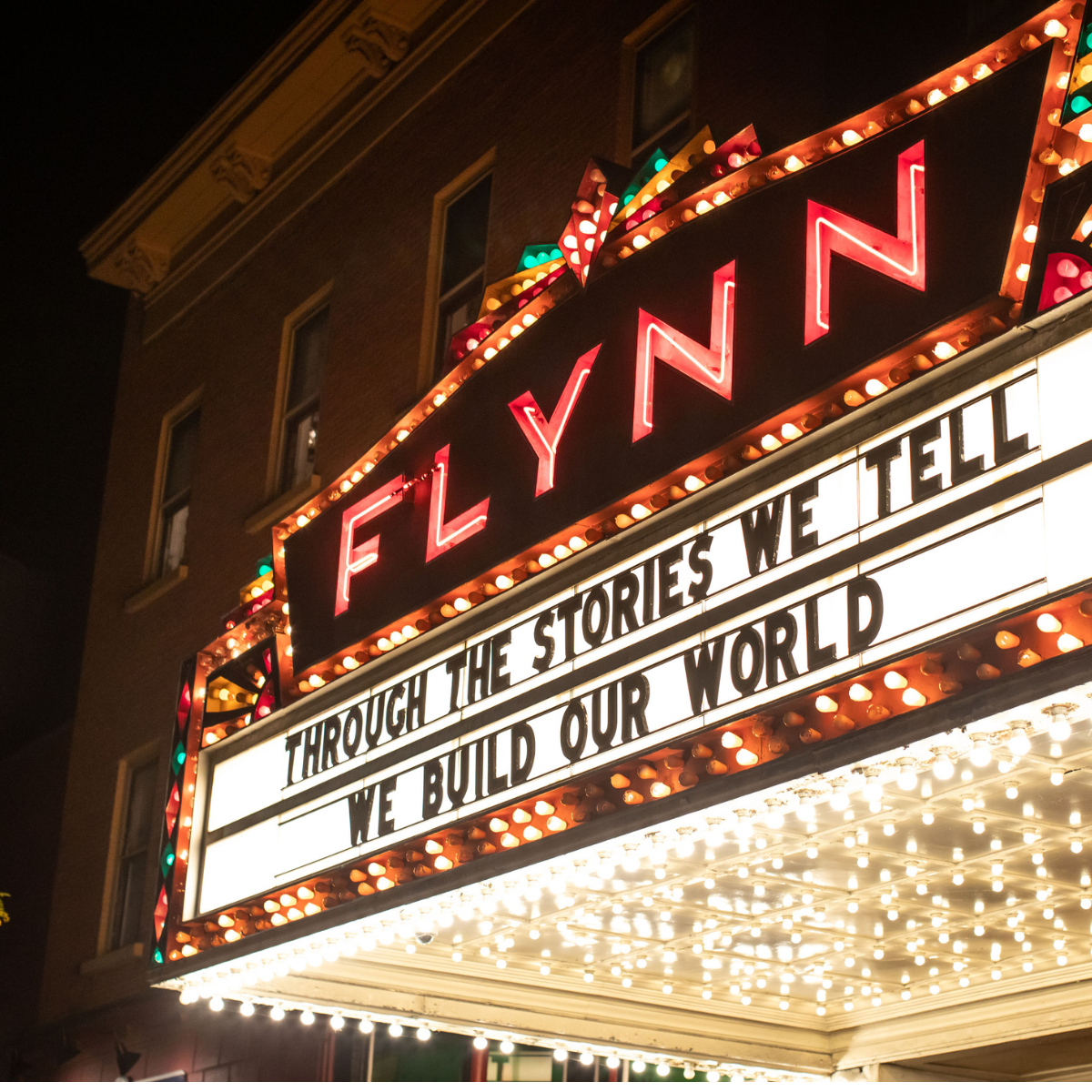 Marquee sign for the Flynn Theater with the message, 'Through the stories we tell, we build our world,' illuminated by bright lights at night.