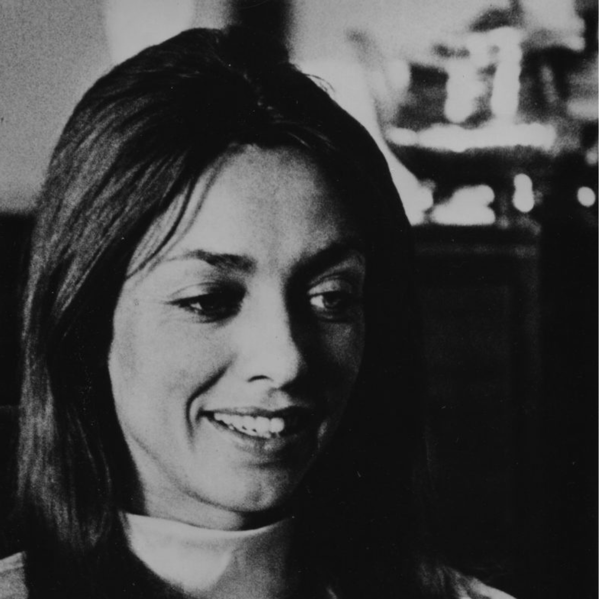 A black and white photo of a woman smiling with long hair, wearing a light-colored top, with a blurred background.