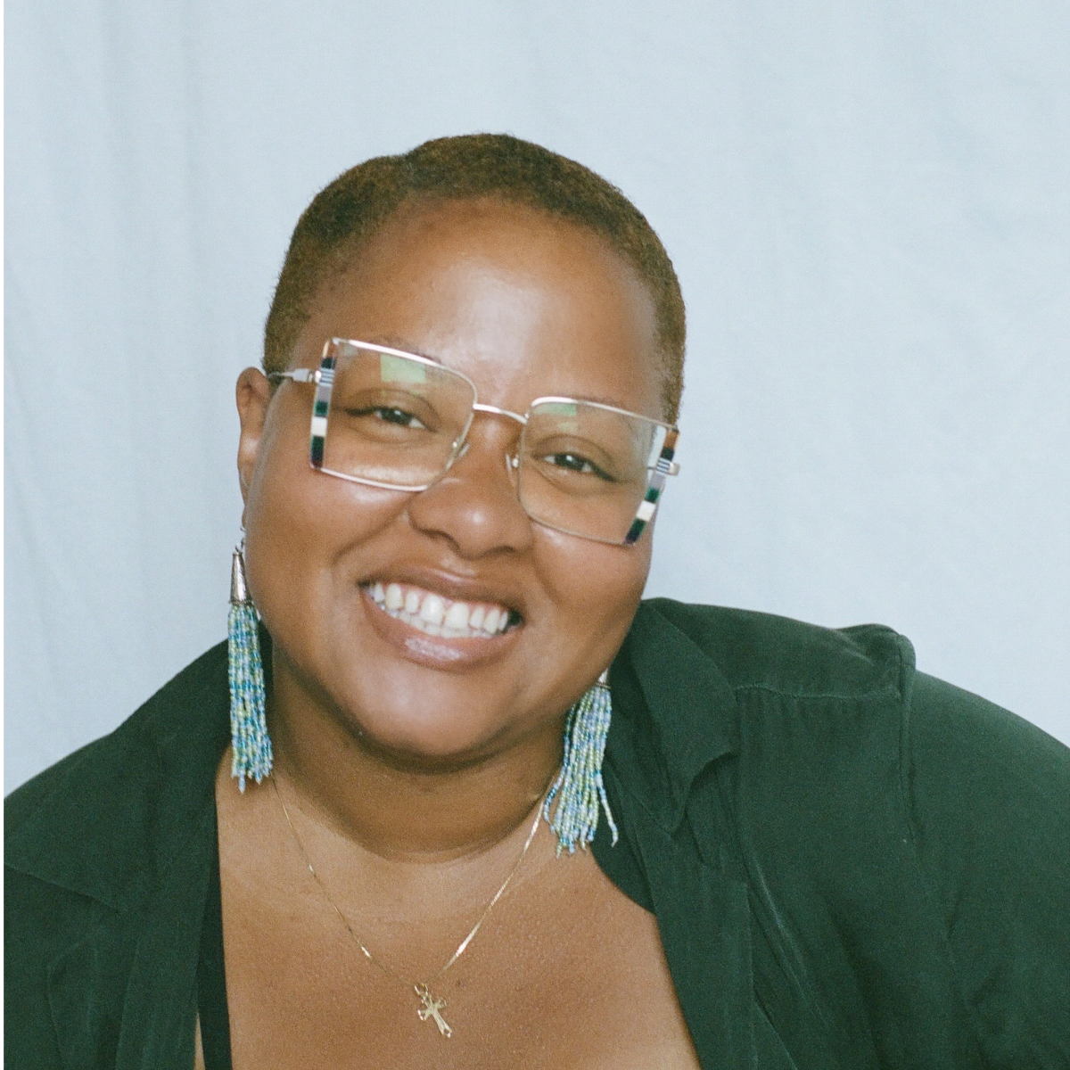 A woman with short hair, wearing large glasses, colorful beaded earrings, and a cross necklace, smiling at the camera against a plain background.