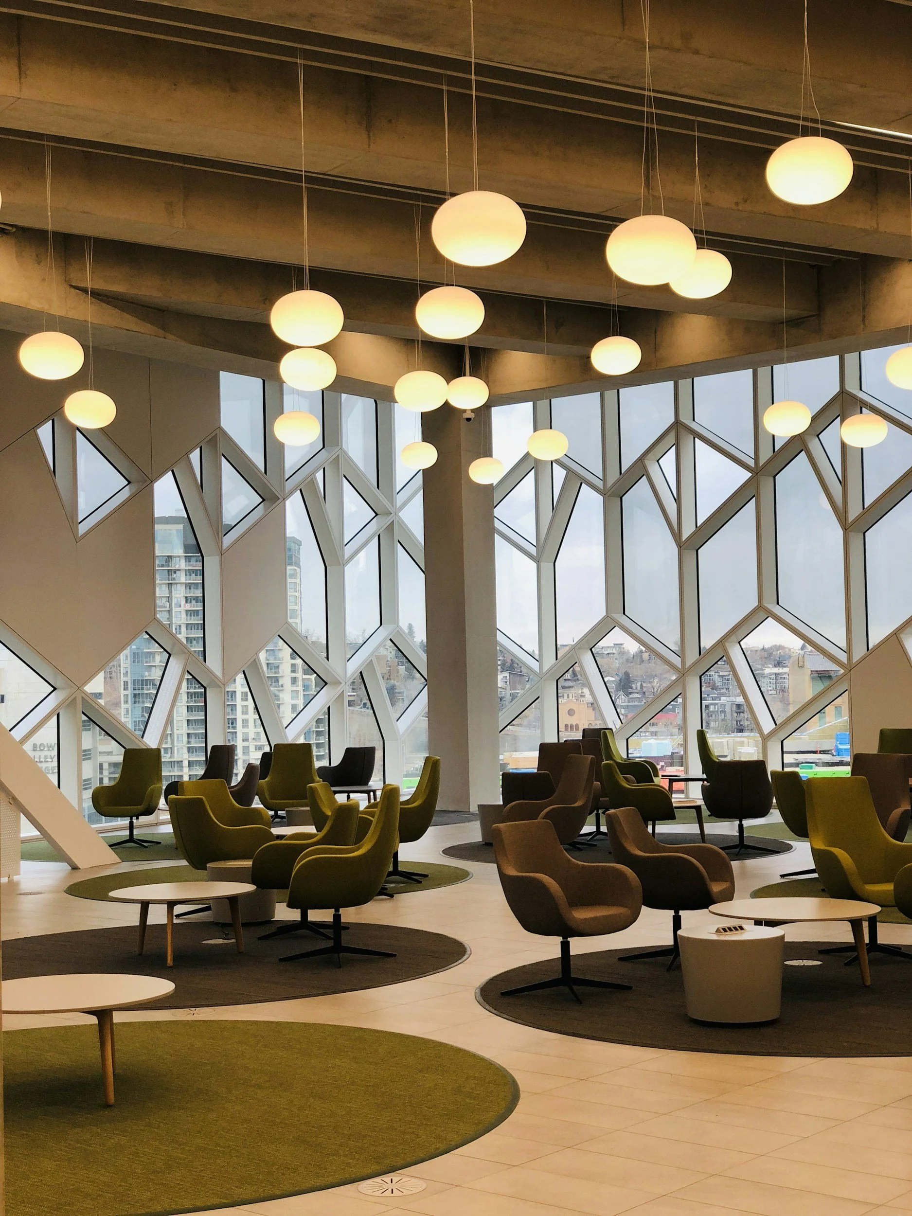 Modern lobby or lounge area with large windows and geometric wall design, furnished with green and brown chairs, small tables, and circular rugs.