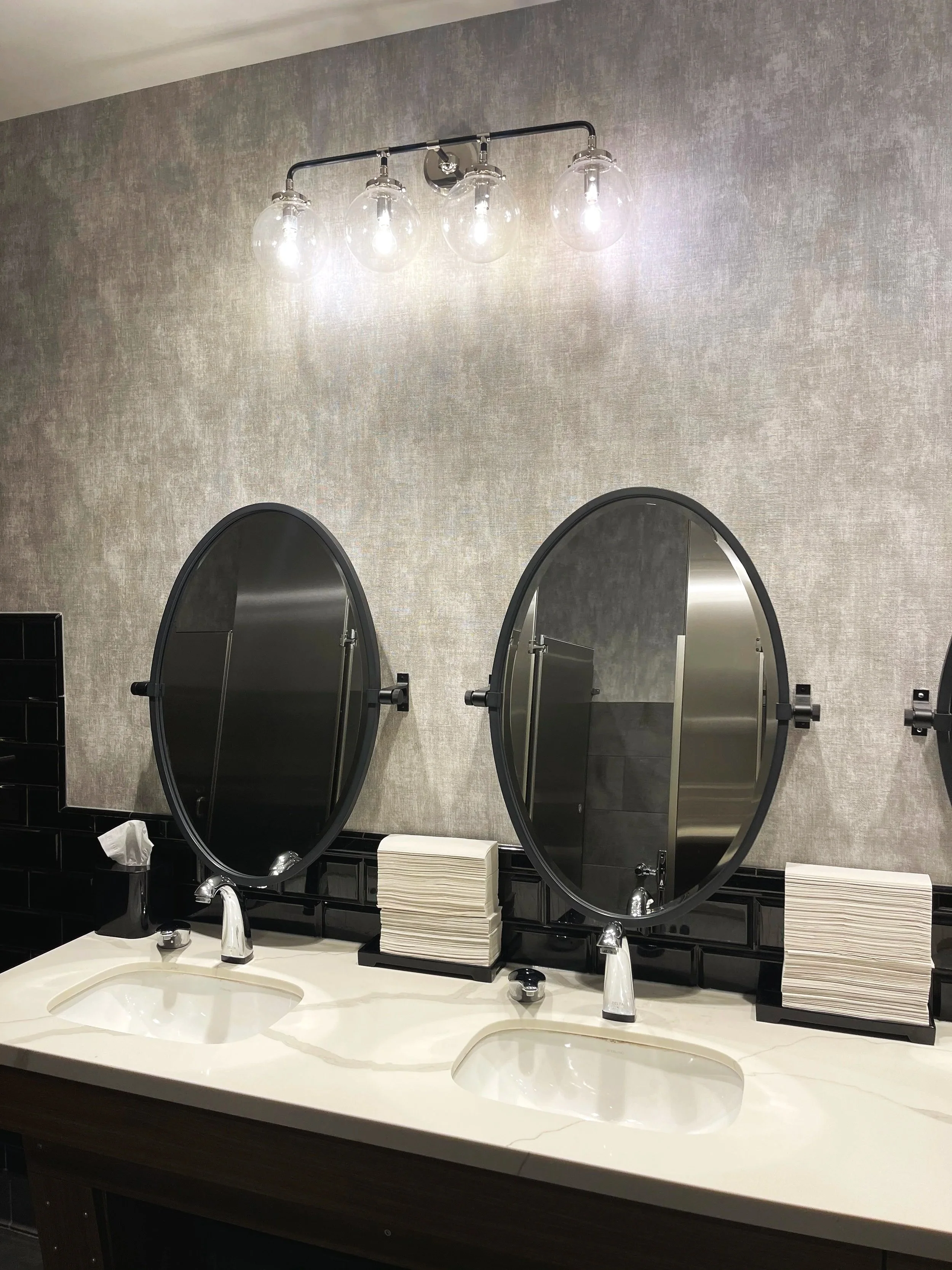 Two bathroom sinks with faucets, a mirror, a stack of paper towels, and a black wall-mounted light fixture with four glass globes, set against a light textured wall.