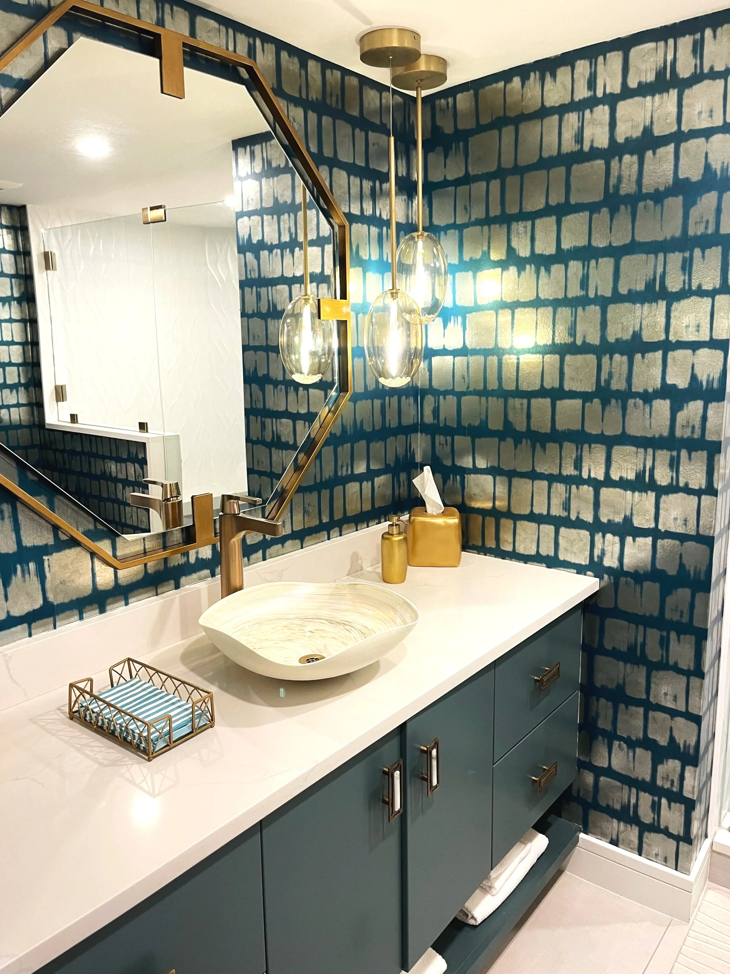 A modern bathroom vanity with a white countertop, a textured oval sink, and a gold soap dispenser. There is a gold and glass tissue box holder and a small decorative basket with striped fabric. A large mirror with a geometric gold frame reflects a se