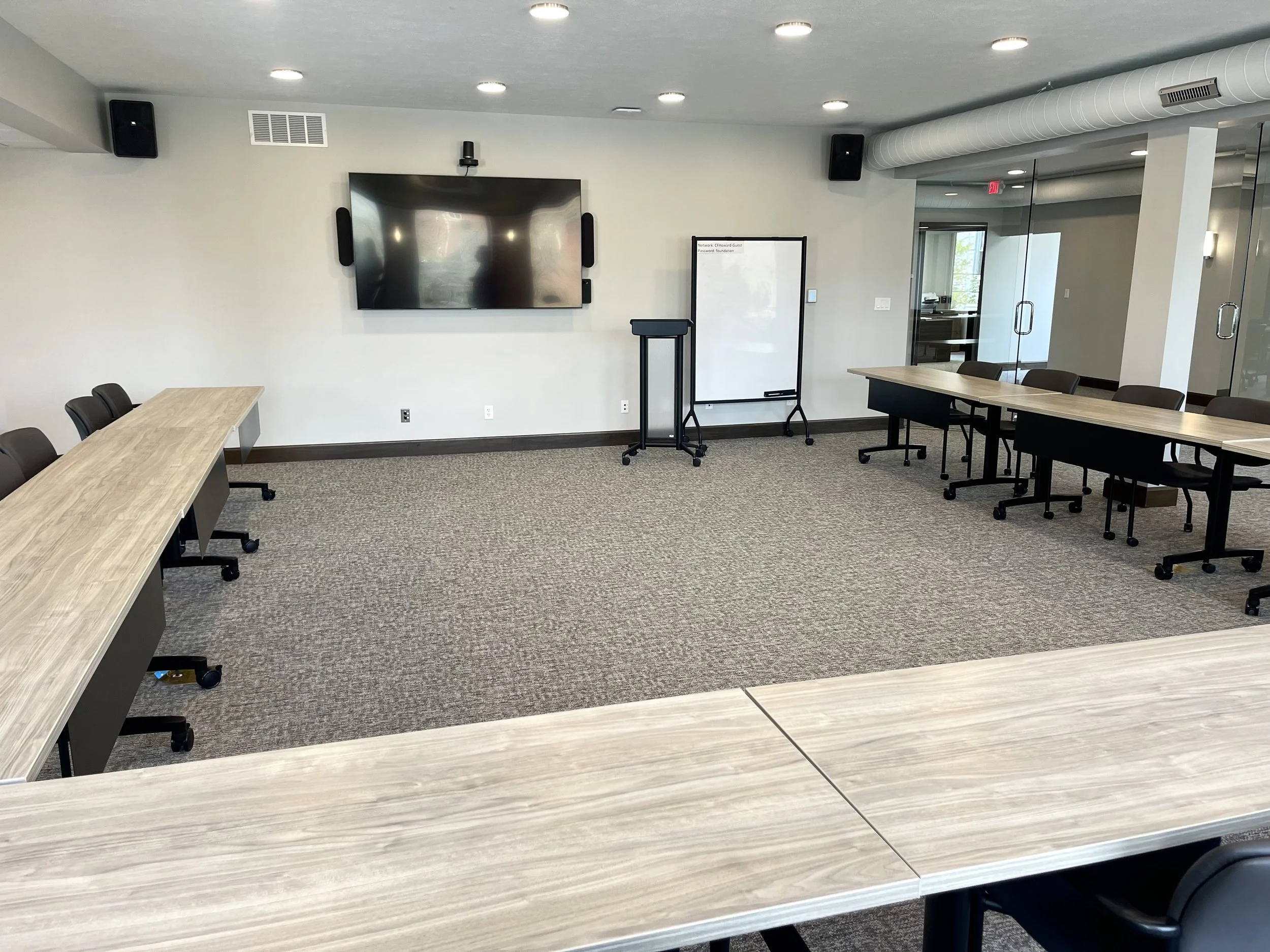 Empty conference or meeting room with a large flat screen TV, whiteboard on a mobile stand, and chairs around a long table.