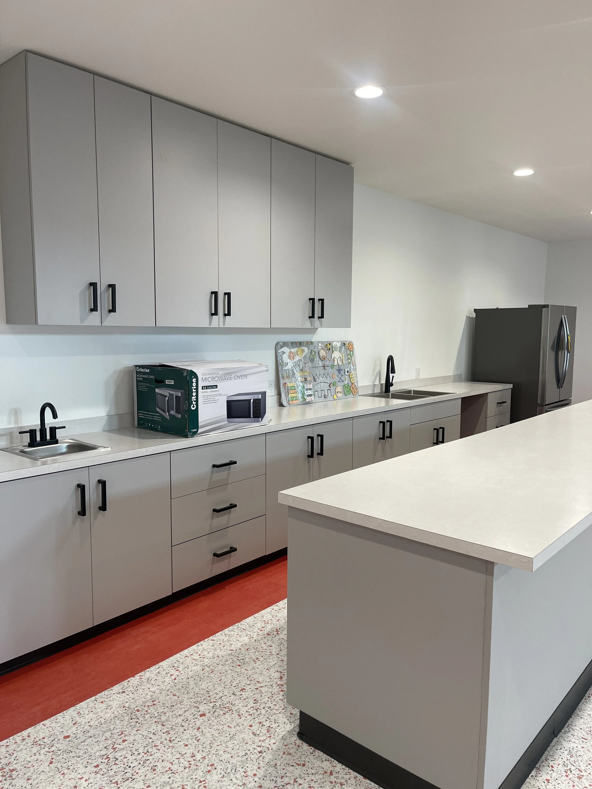 Modern kitchen with gray cabinets, white countertops, a black faucet, a microwave box, a cutting board with kitchen utensils, a black refrigerator, and red and speckled flooring.