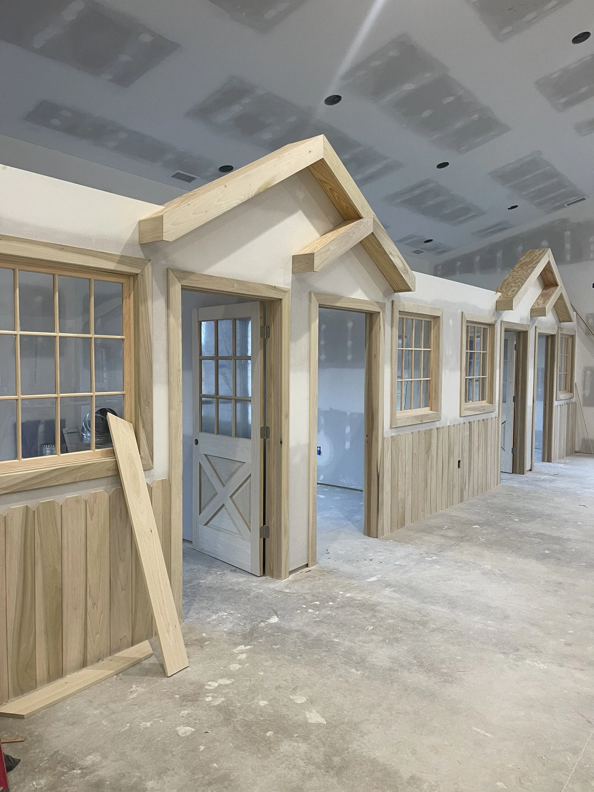 Interior of a building under construction with unfinished walls, multiple small house-shaped wooden structures with windows, and a concrete floor.